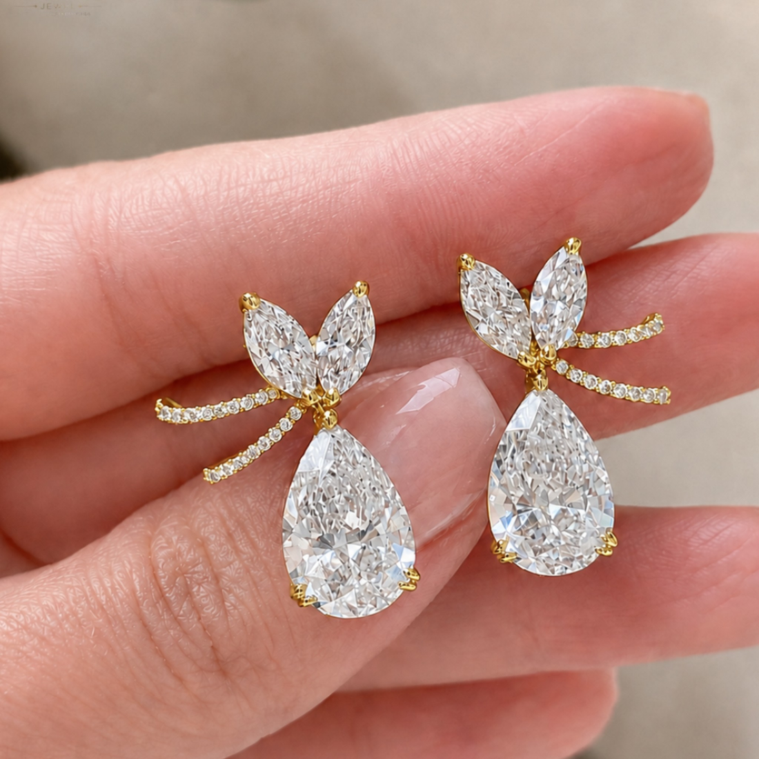 A pair of gold and diamond drop earrings held on a person's fingers. Each earring features a teardrop-shaped clear gemstone with smaller marquise and round-cut clear stones in a yellow gold setting.