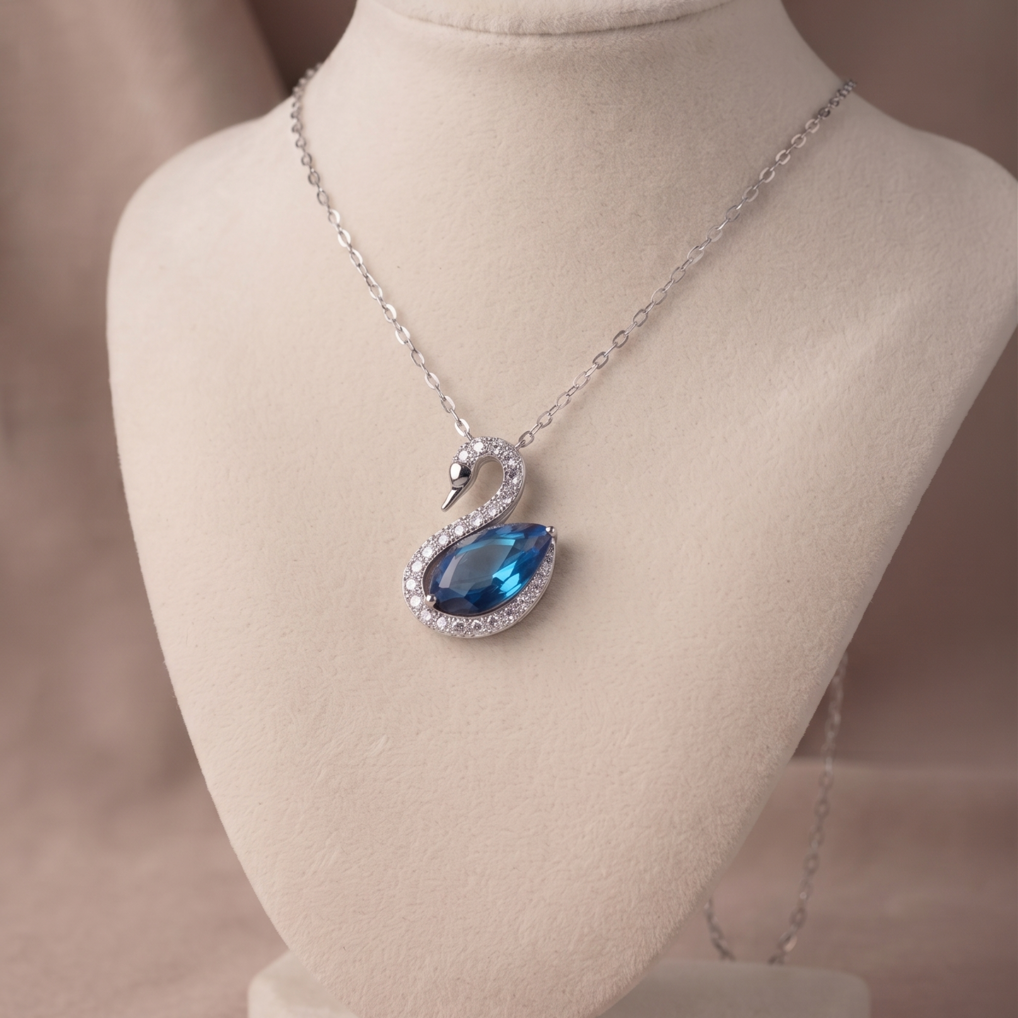 A silver-tone swan pendant necklace with a blue teardrop gemstone and clear crystal accents, displayed on a beige jewelry bust.
