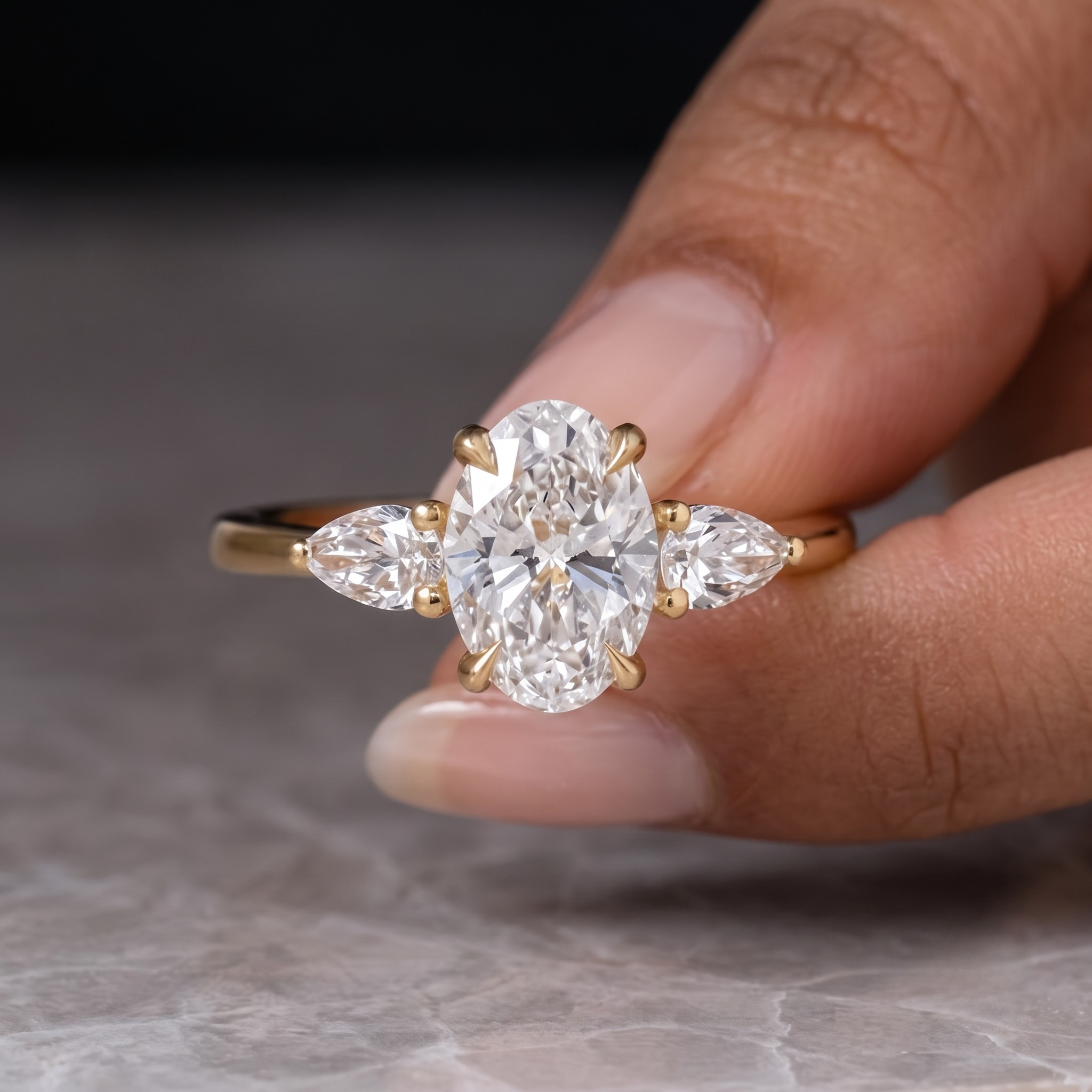 A yellow gold ring with an oval center stone and two marquise-cut side stones, held between fingers against a soft gray marble surface.