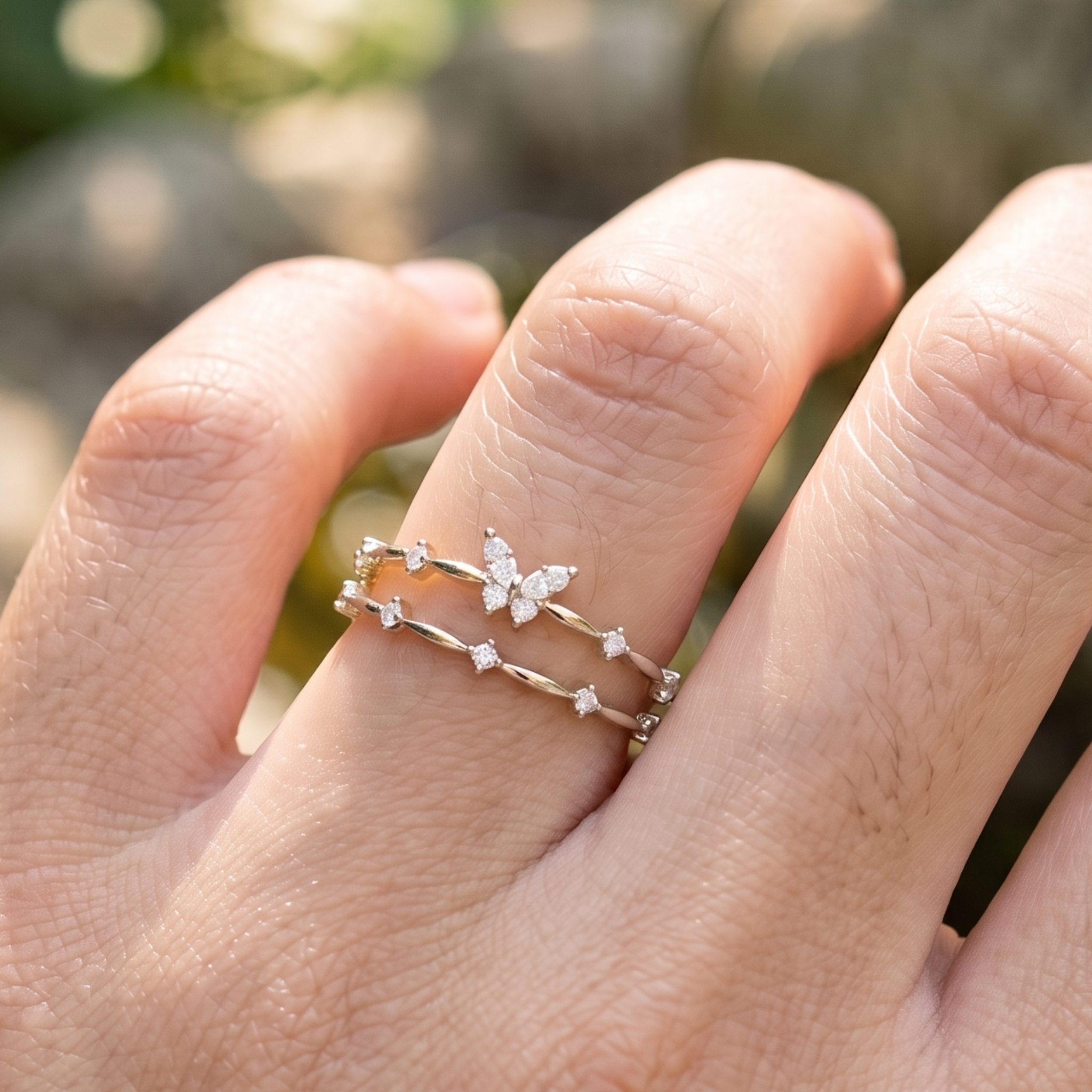 A close-up of a hand wearing two delicate silver rings. One ring features a small butterfly motif set with clear stones, and the other is a thin silver band with small star-shaped clear stone accents, set against a softly blurred outdoor background.