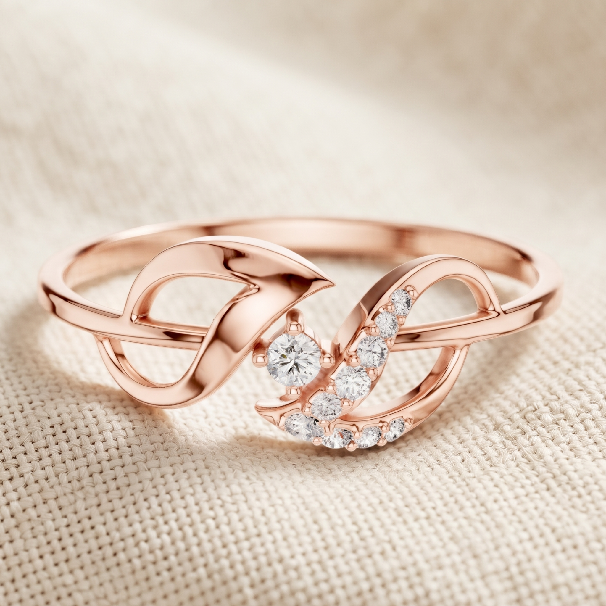 A rose gold ring with a stylized infinity symbol design, featuring a central round diamond and a curved row of small clear stones, resting on a textured beige fabric background.