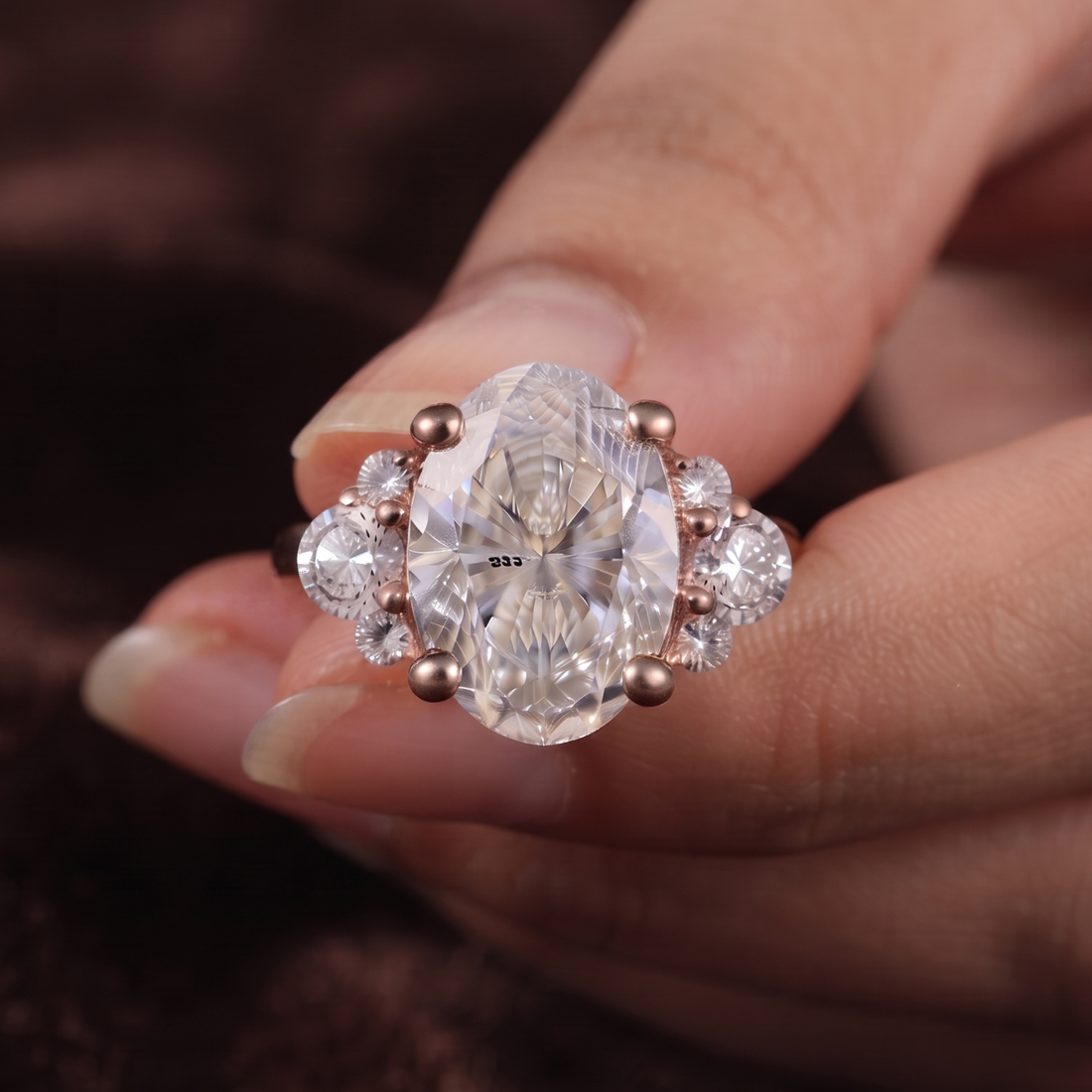 A close-up of a hand wearing a rose gold ring with a large oval clear gemstone and smaller accent stones, set against a dark brown textured background.
