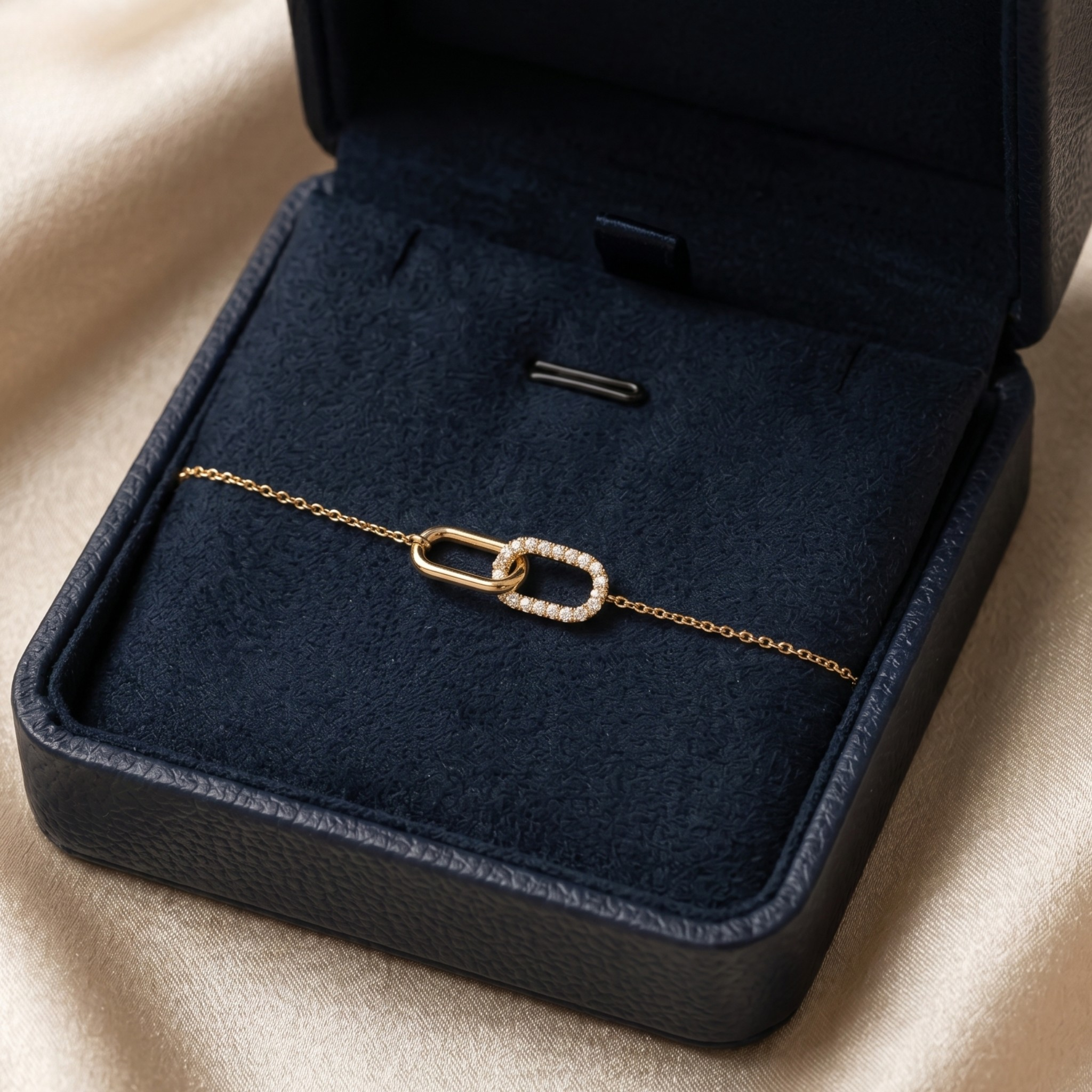 A delicate gold chain bracelet with two interlocking oval links, one adorned with small clear stones, displayed inside an open dark navy jewelry box on a beige fabric background.