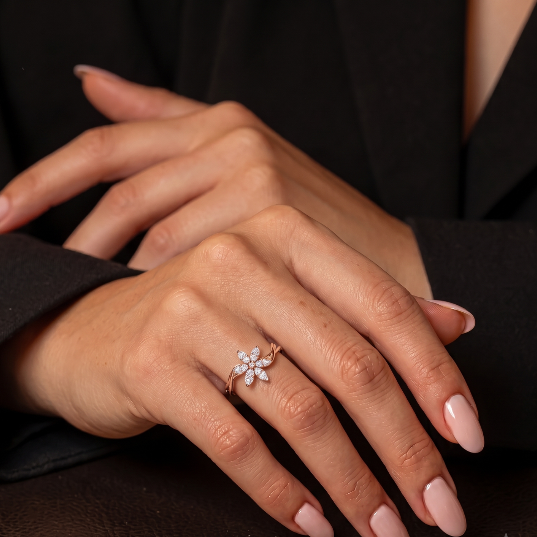 A close-up of a person's hands wearing a delicate rose gold ring with a small flower-shaped cluster of clear stones. The person is wearing a black blazer, and their nails are painted a soft pink.