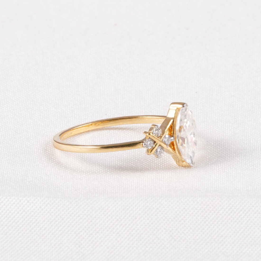 A yellow gold ring featuring a marquise-cut clear gemstone in a prong setting with small accent diamonds, shown on a textured white background.