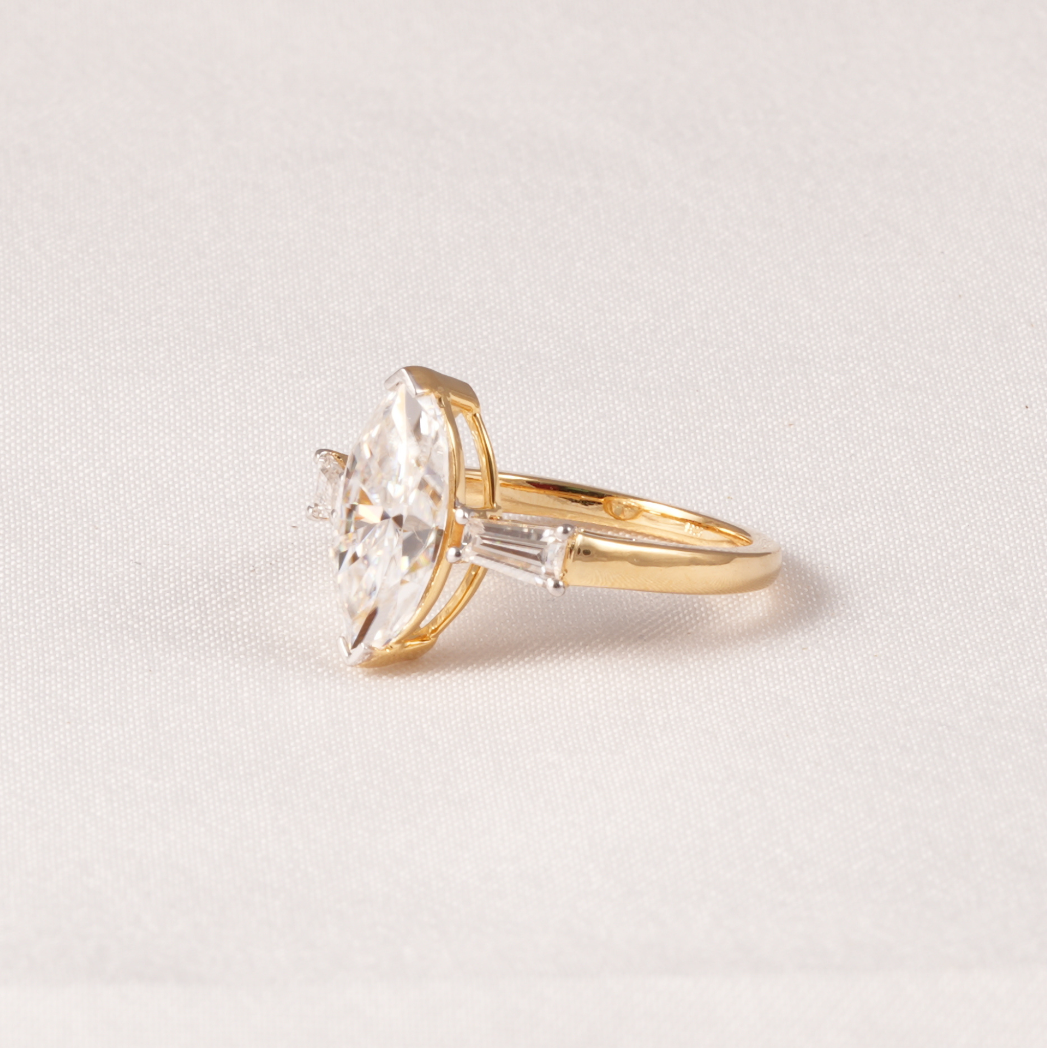 A yellow gold ring featuring a large marquise-cut clear gemstone and smaller side stones, photographed on a light neutral fabric background.