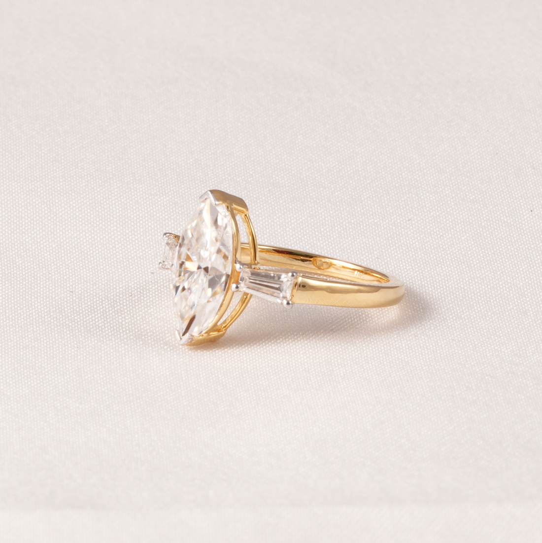 A yellow gold ring featuring a large marquise-cut clear gemstone and smaller side stones, photographed on a light neutral fabric background.