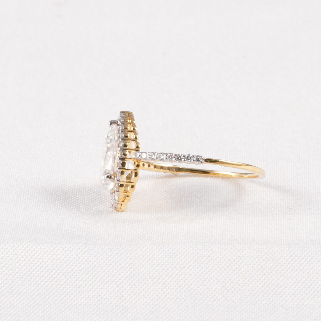 A yellow gold ring with a halo setting and a pavé band, featuring a clear round-cut center stone and small clear accent stones, shown on a white textured background.