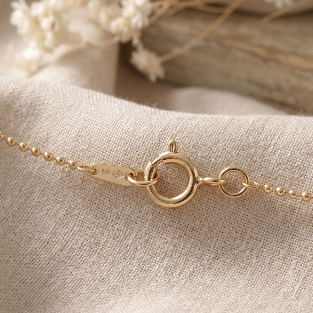 Close-up of a delicate gold chain bracelet with a lobster clasp, resting on a textured beige linen fabric with dried white flowers in the background.