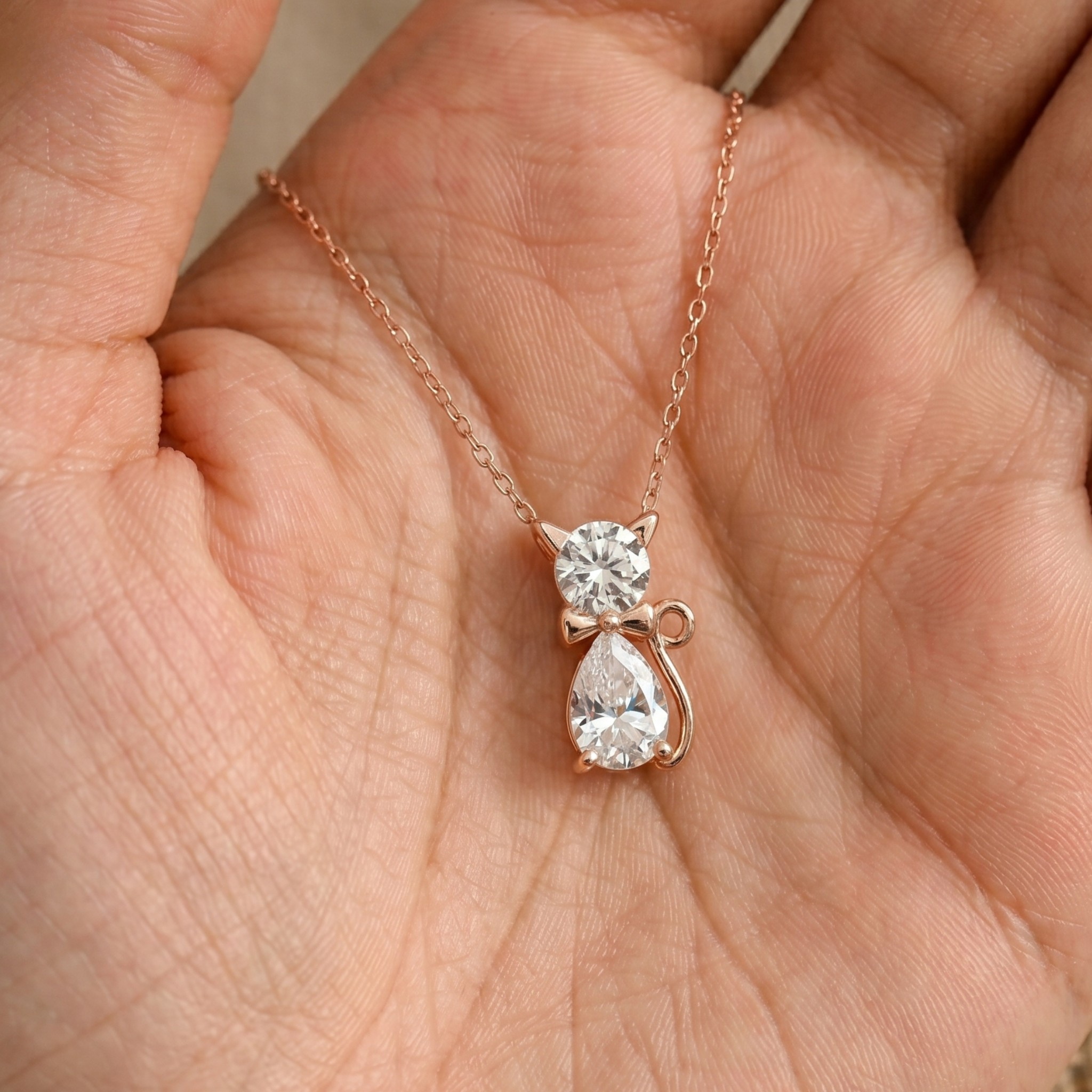 A rose gold necklace with a cat-shaped pendant featuring a round clear gemstone and a teardrop-shaped clear gemstone, displayed on an open hand.