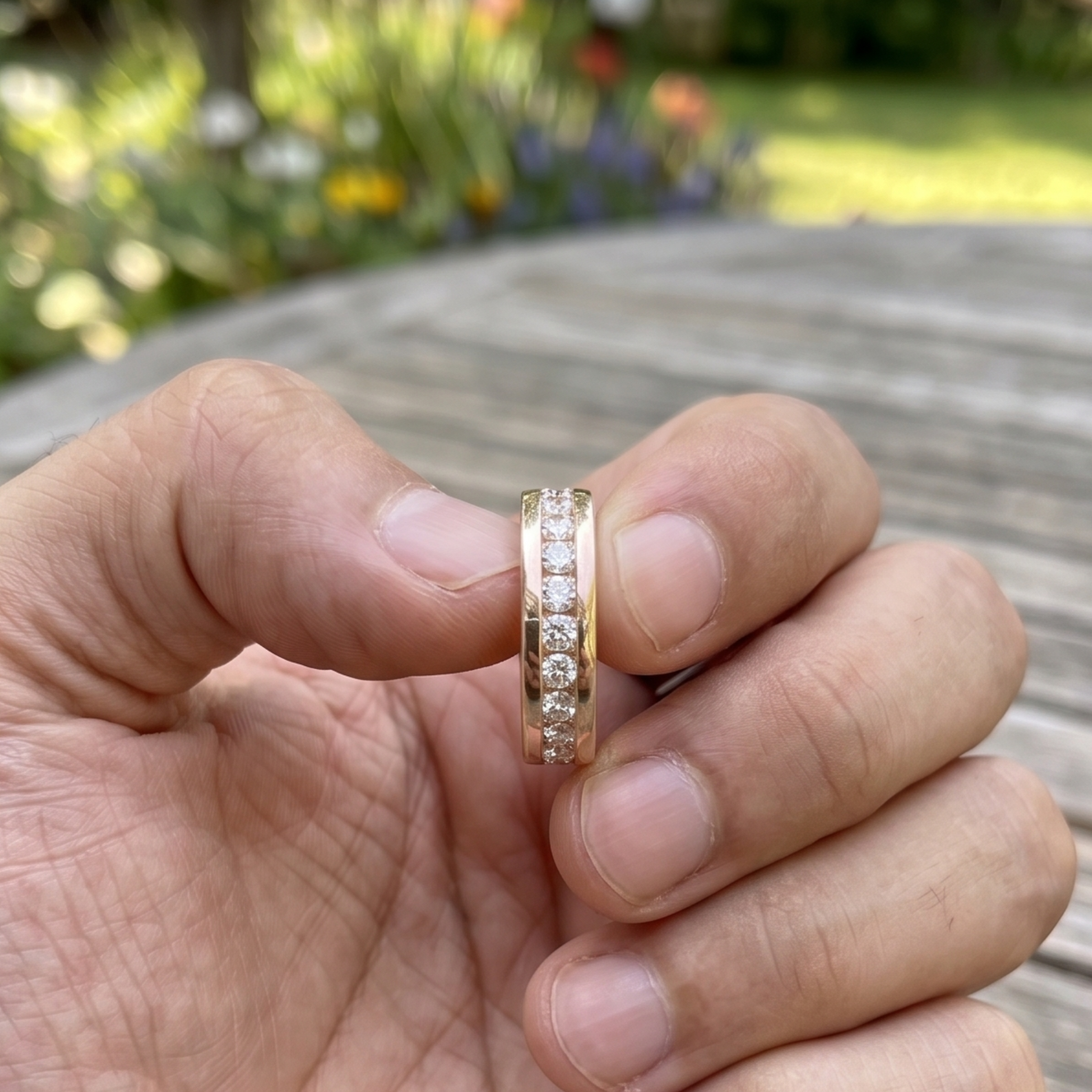 A gold band ring with a row of clear stones, held between fingers outdoors with a blurred garden and wooden table in the background.