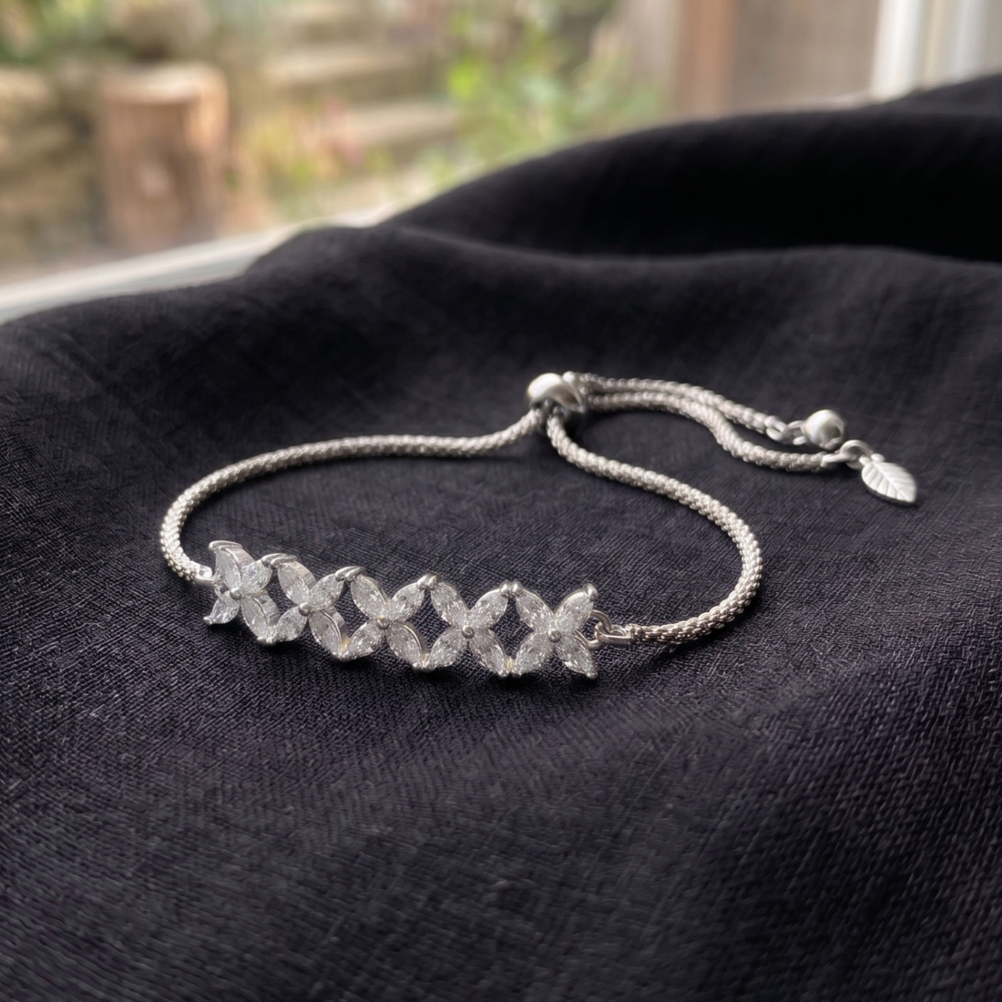 A silver-toned adjustable bracelet with a row of four interlocking flower motifs, displayed on a dark textured fabric surface.