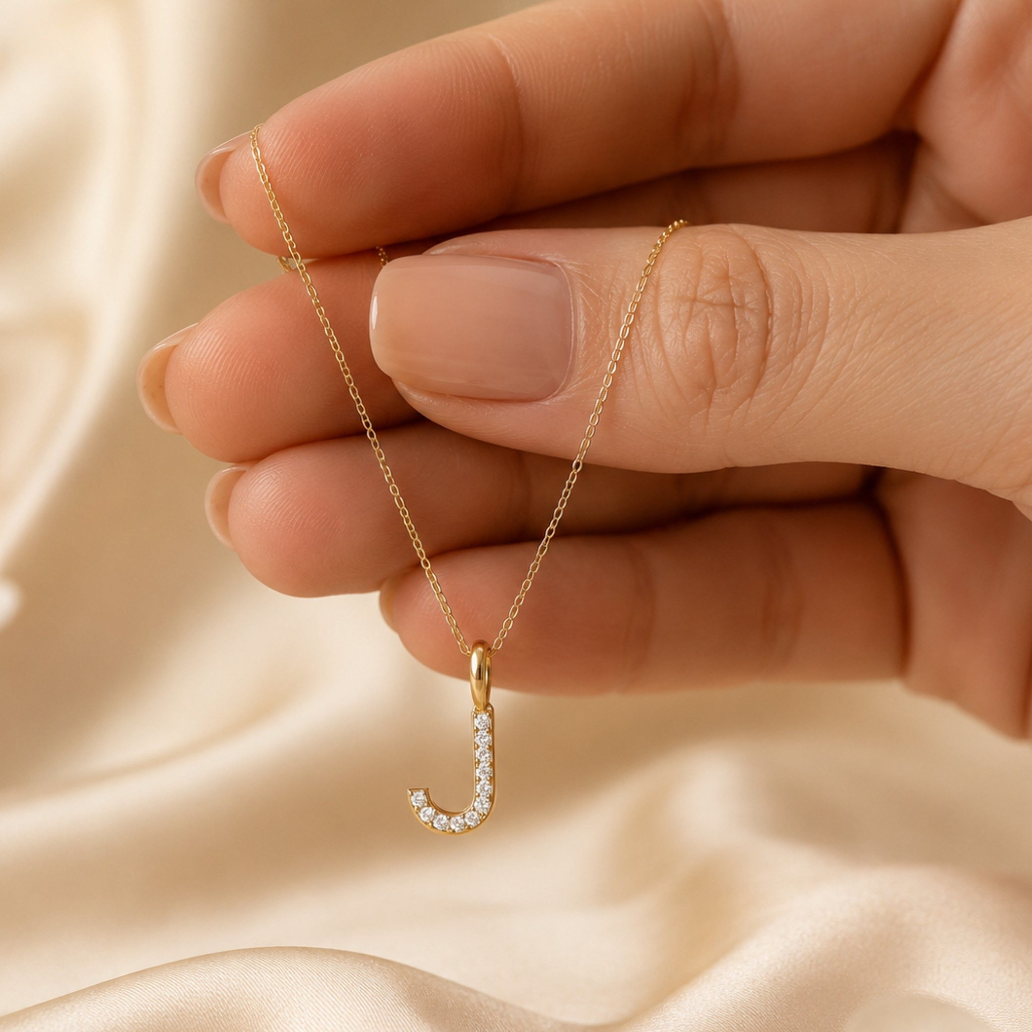 A close-up of a hand holding a delicate gold chain necklace with a small gold initial pendant set with clear stones, against a soft beige satin background.