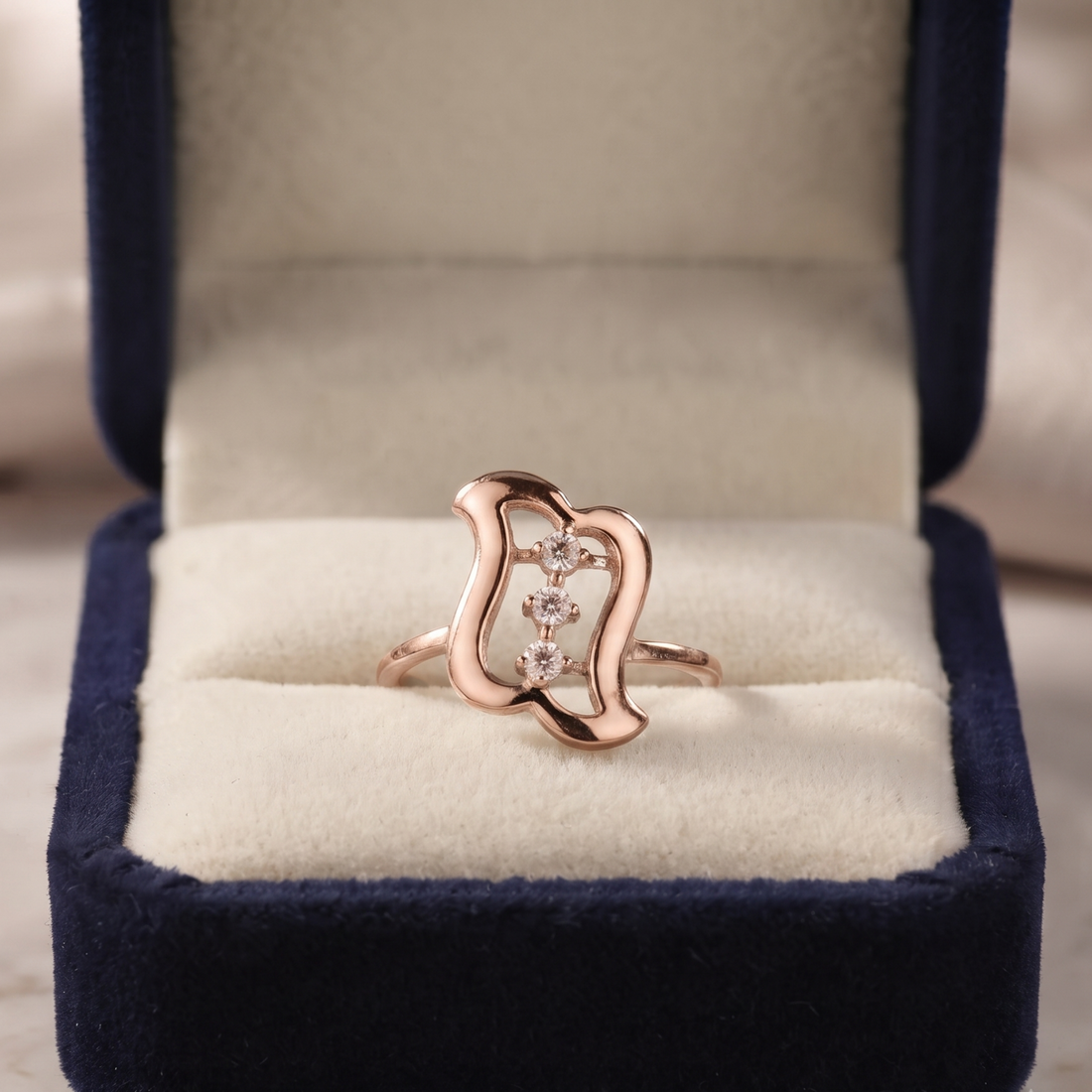 A rose gold ring with a wavy openwork design and three small clear stones, displayed in an open navy blue velvet ring box with a cream cushion.