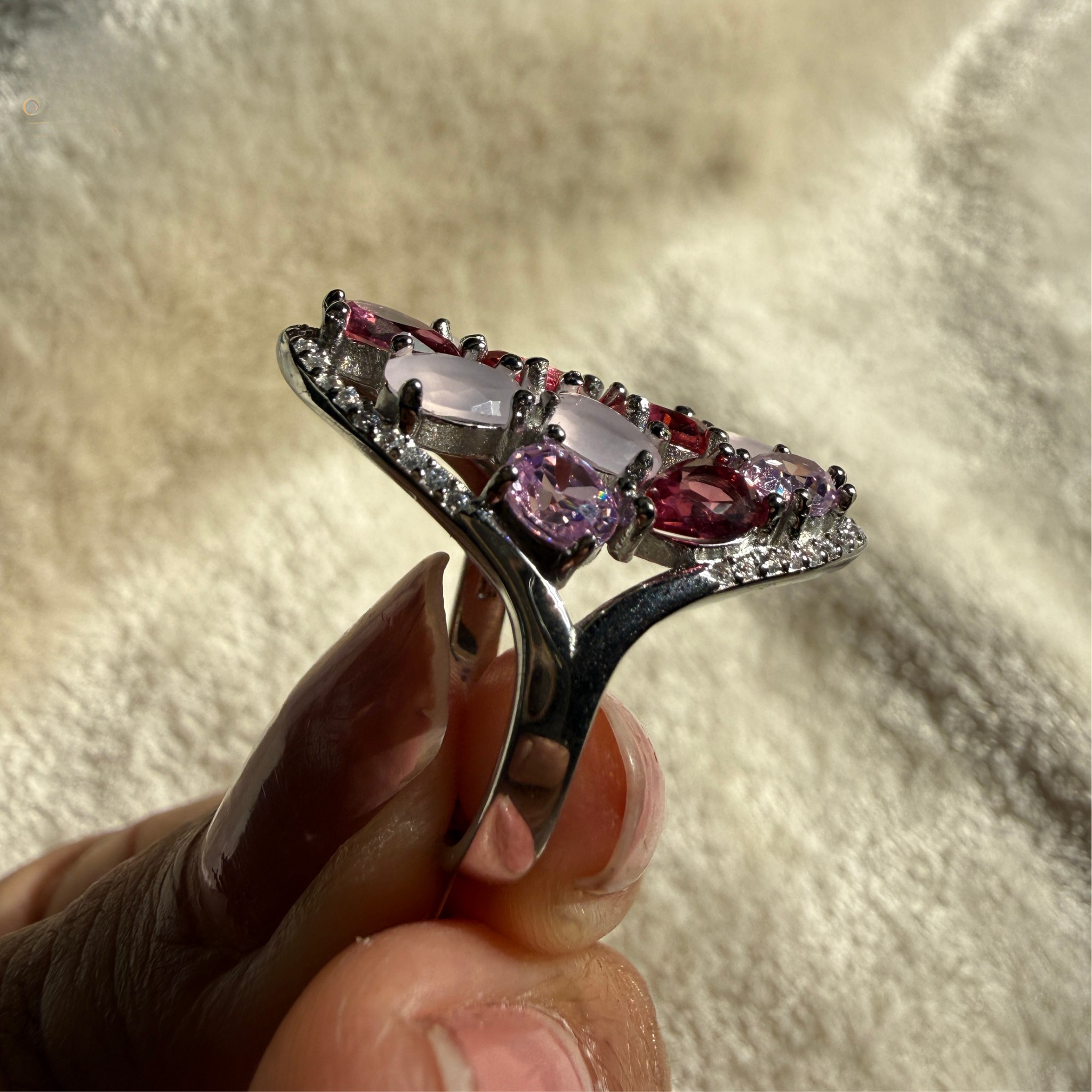 A silver ring with a curved band and a row of pink gemstones, held between fingers against a soft beige textured background.