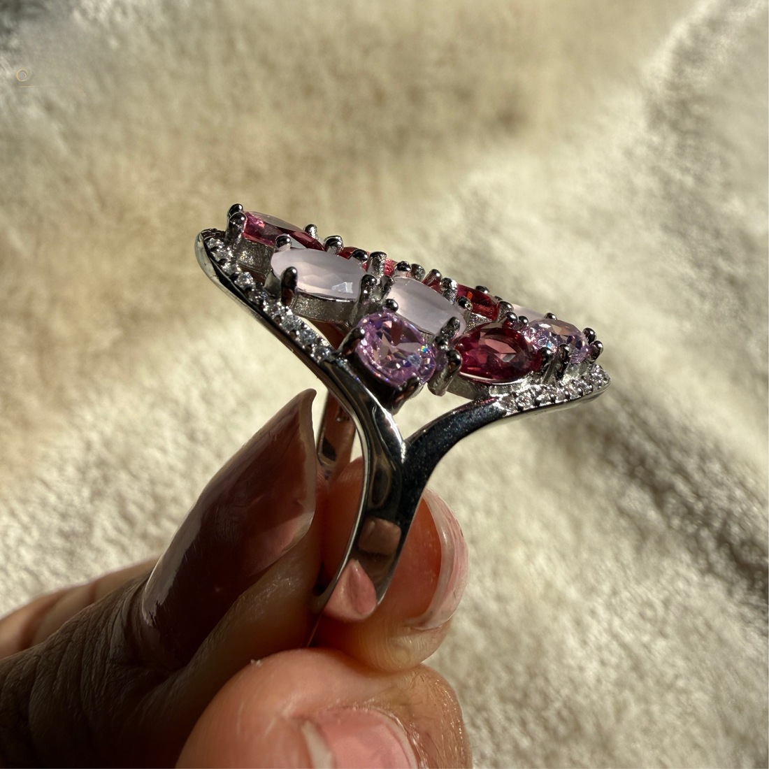 A silver ring with a curved band and a row of pink gemstones, held between fingers against a soft beige textured background.