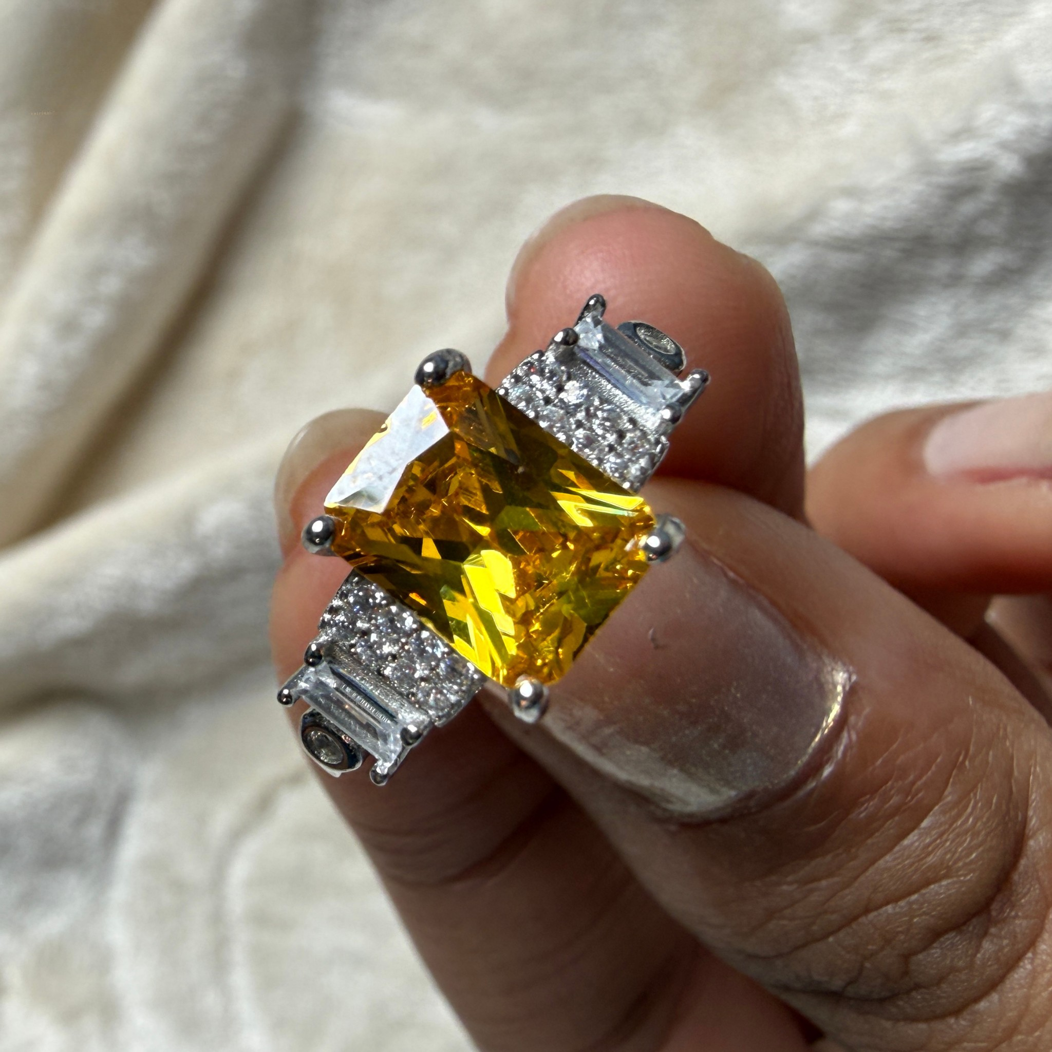 A close-up of a hand holding a silver ring featuring a large yellow cushion-cut center stone and smaller clear baguette side stones, set against a soft beige fabric background.
