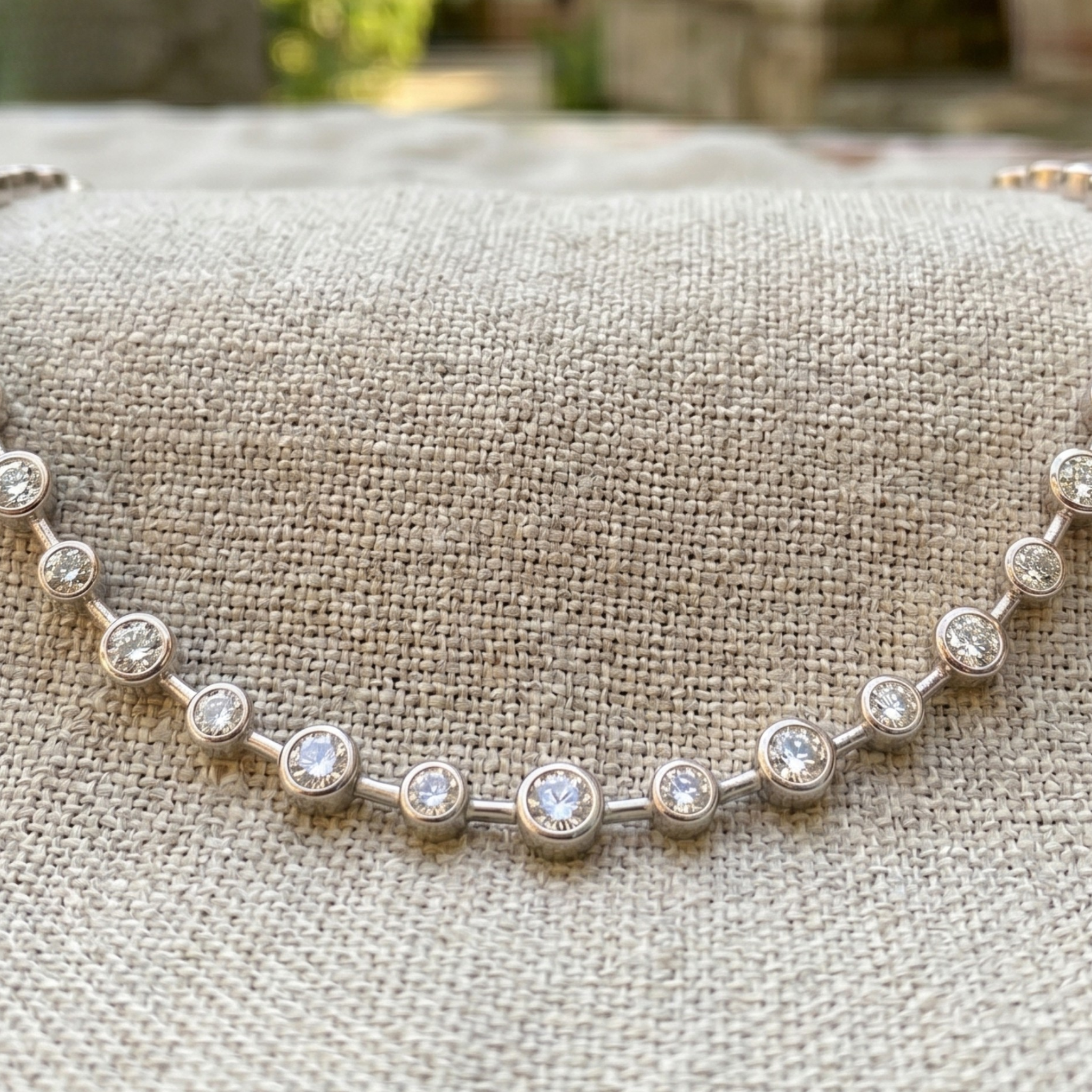 A silver-toned tennis bracelet with round bezel-set clear stones, displayed on a textured beige linen fabric.