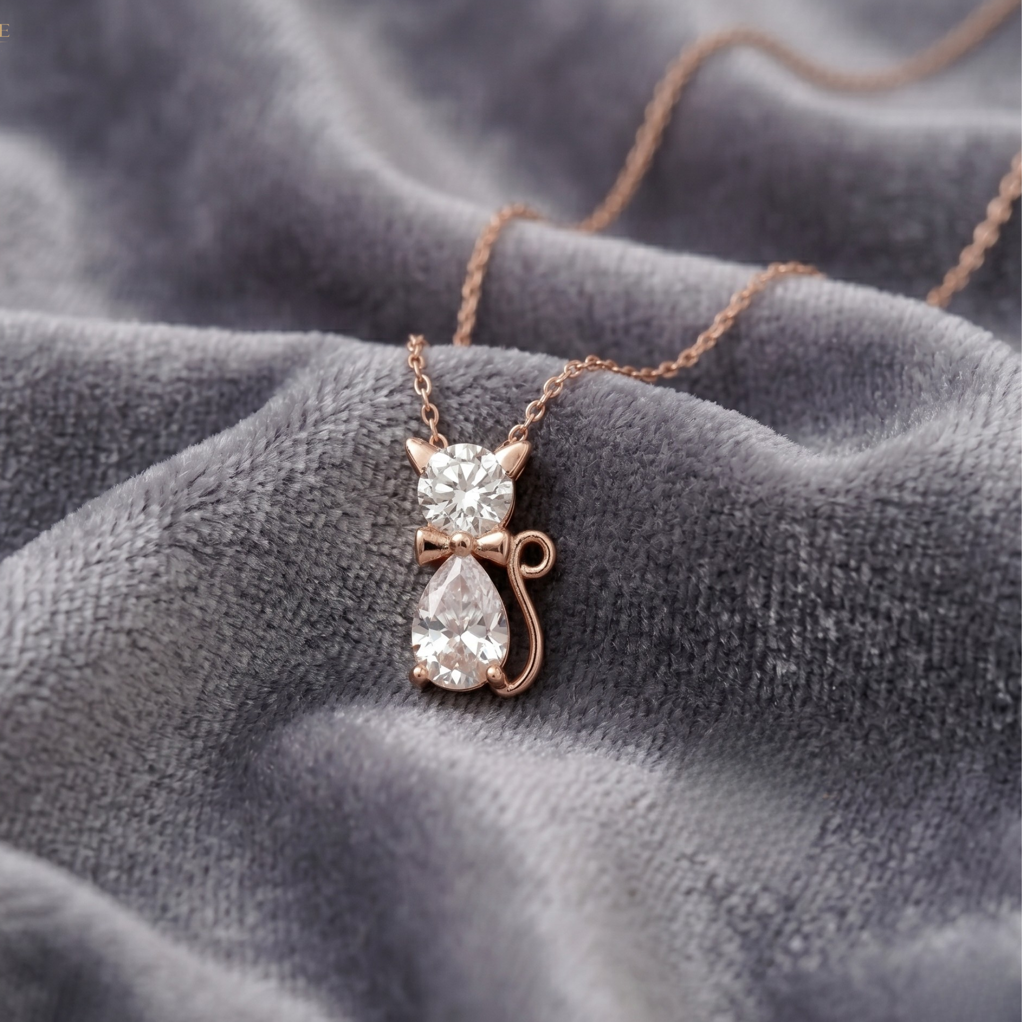 A rose gold cat pendant necklace with a clear teardrop gemstone and a small clear gemstone bow, resting on a textured gray velvet fabric.