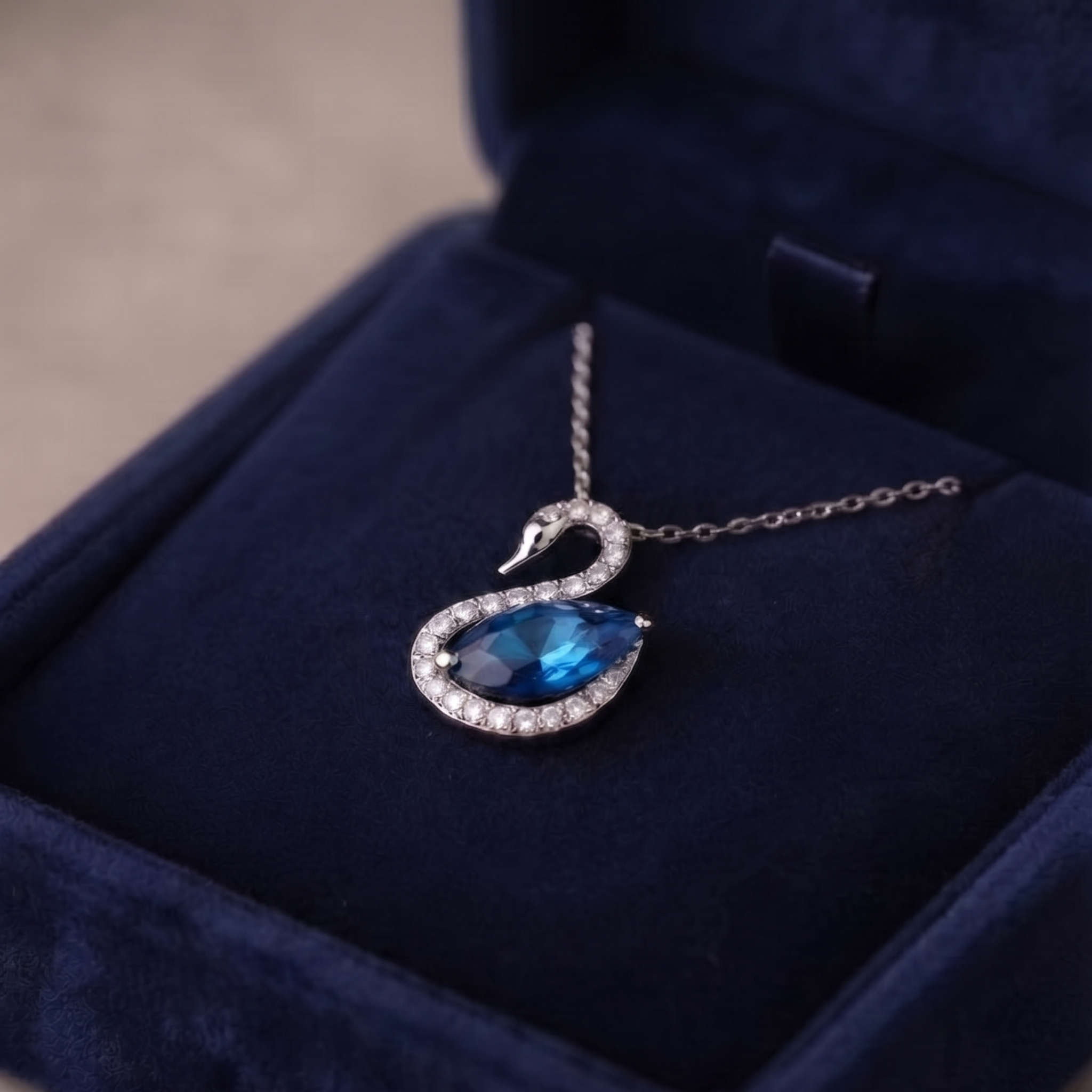 A silver-toned swan pendant necklace with a blue teardrop gemstone and small clear stones, displayed inside a dark blue velvet jewelry box.