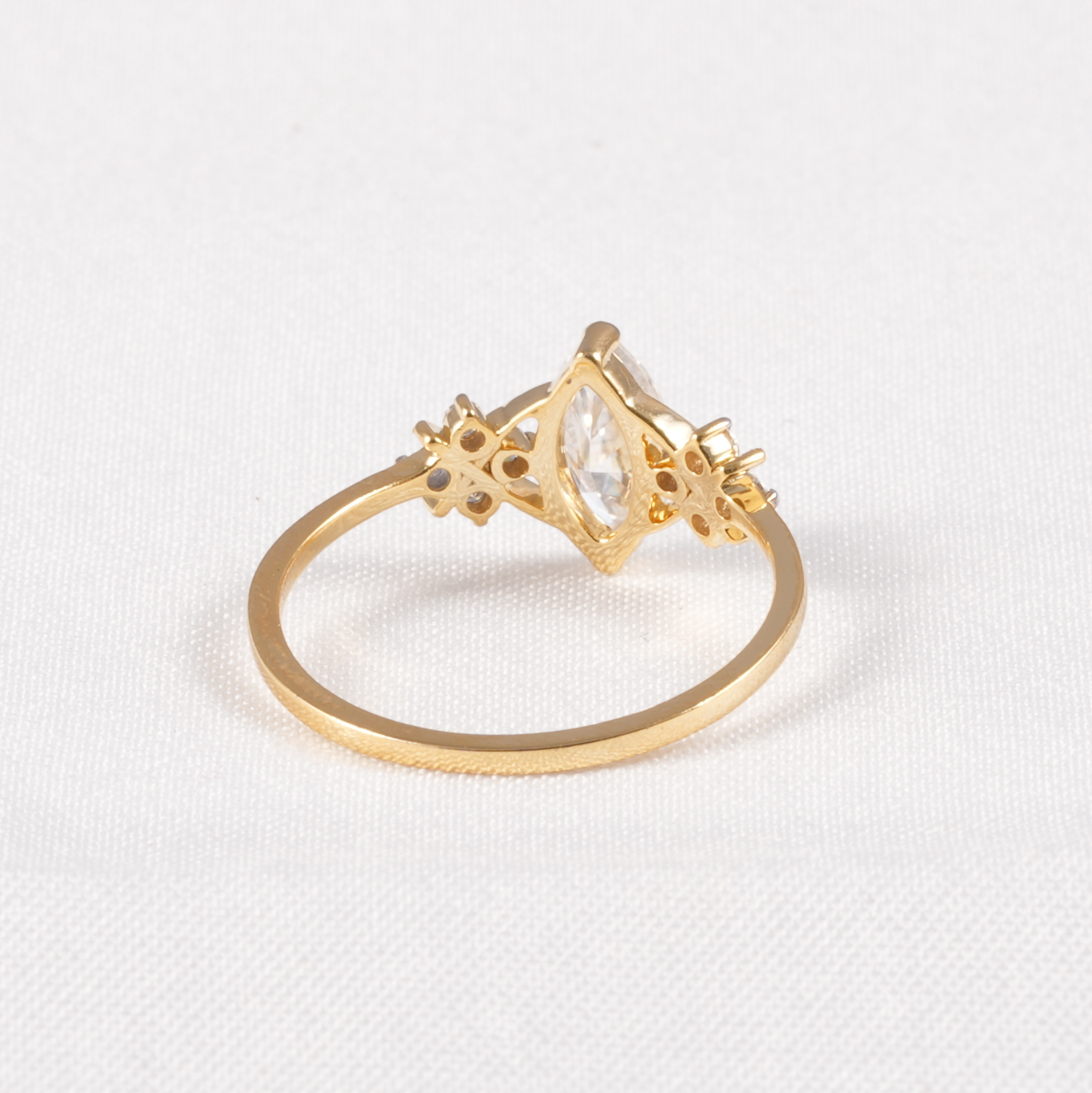 A gold ring with a marquise-cut clear gemstone in a diamond-shaped setting, accented with small round and star-shaped clear stones, shown on a textured white background.