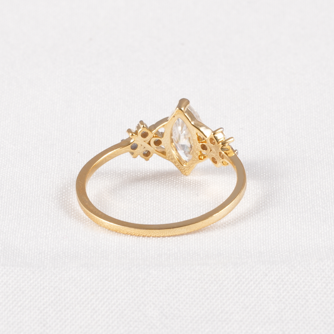 A gold ring with a marquise-cut clear gemstone in a diamond-shaped setting, accented with small round and star-shaped clear stones, shown on a textured white background.
