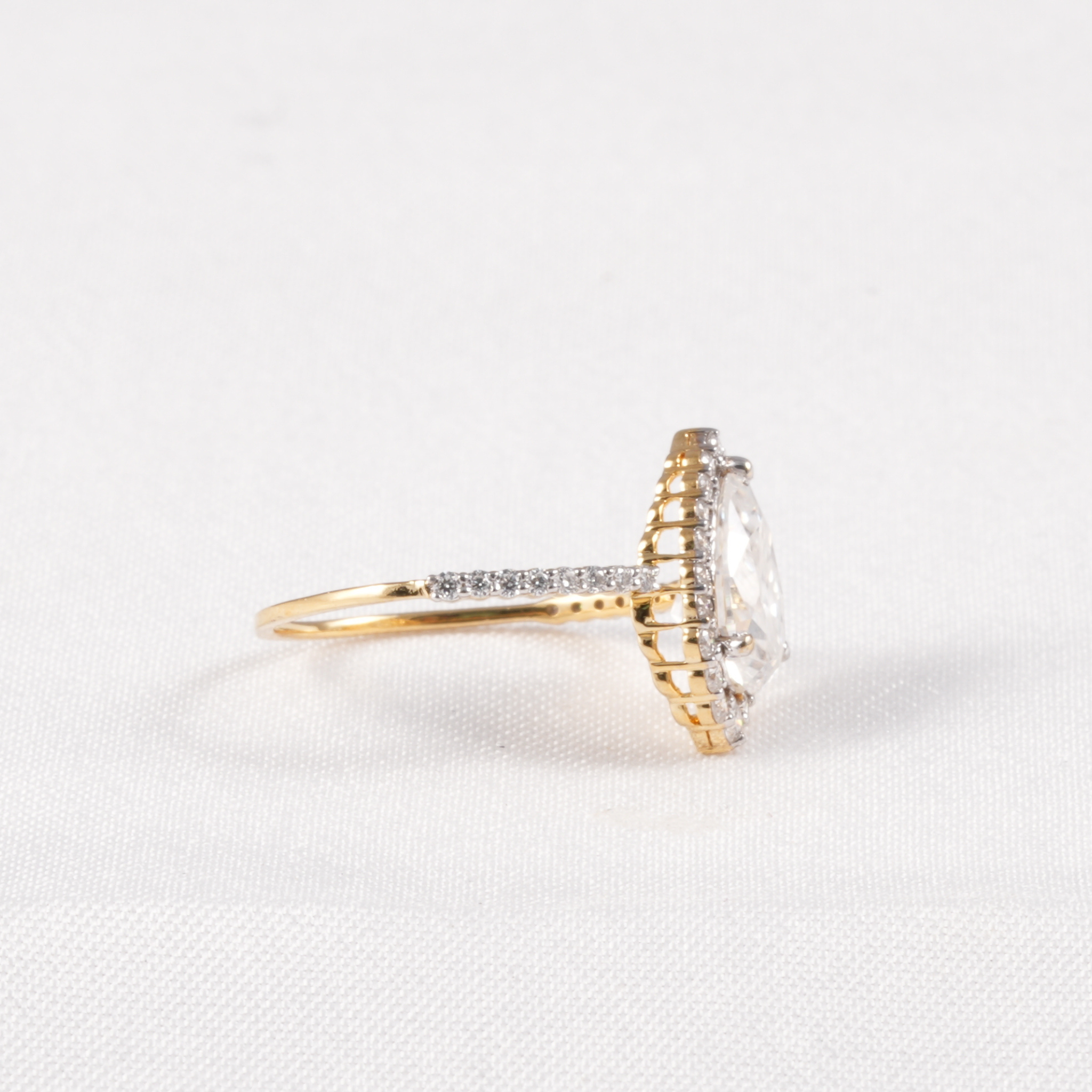 A yellow gold ring featuring a large oval clear gemstone in a halo setting, with a pavé band accented by small diamonds, shown on a textured white background.