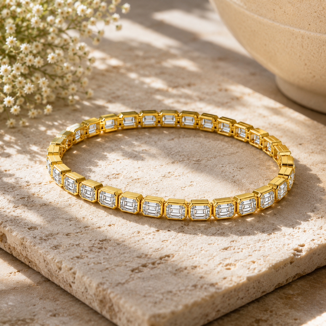 A gold tennis bracelet with rectangular Emerald-cut clear stones, displayed on a textured beige stone slab with soft shadows and small white flowers in the background.