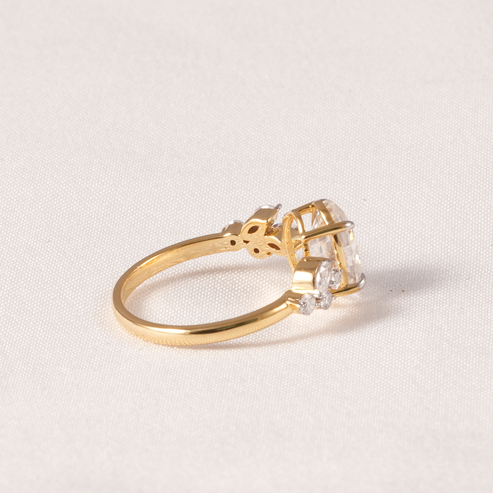 A yellow gold ring featuring a large clear rectangular gemstone in a prong setting, accented by small clear stones on the shoulders, photographed on a light beige textured fabric background.