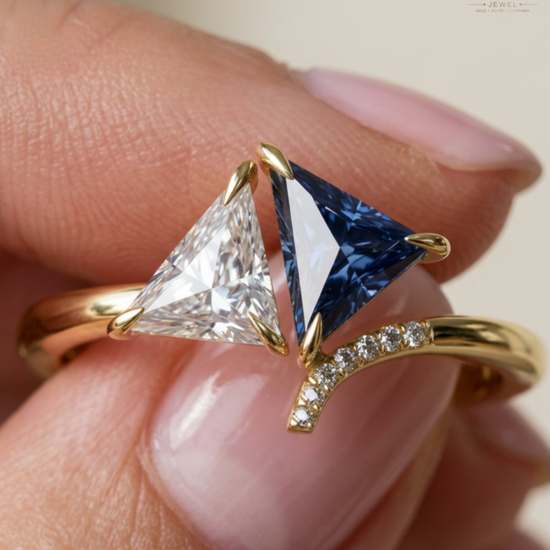 A close-up of a gold ring being worn on a finger, featuring a triangular clear gemstone on one side and a triangular deep blue gemstone on the other, with a curved row of small clear stones on the band.