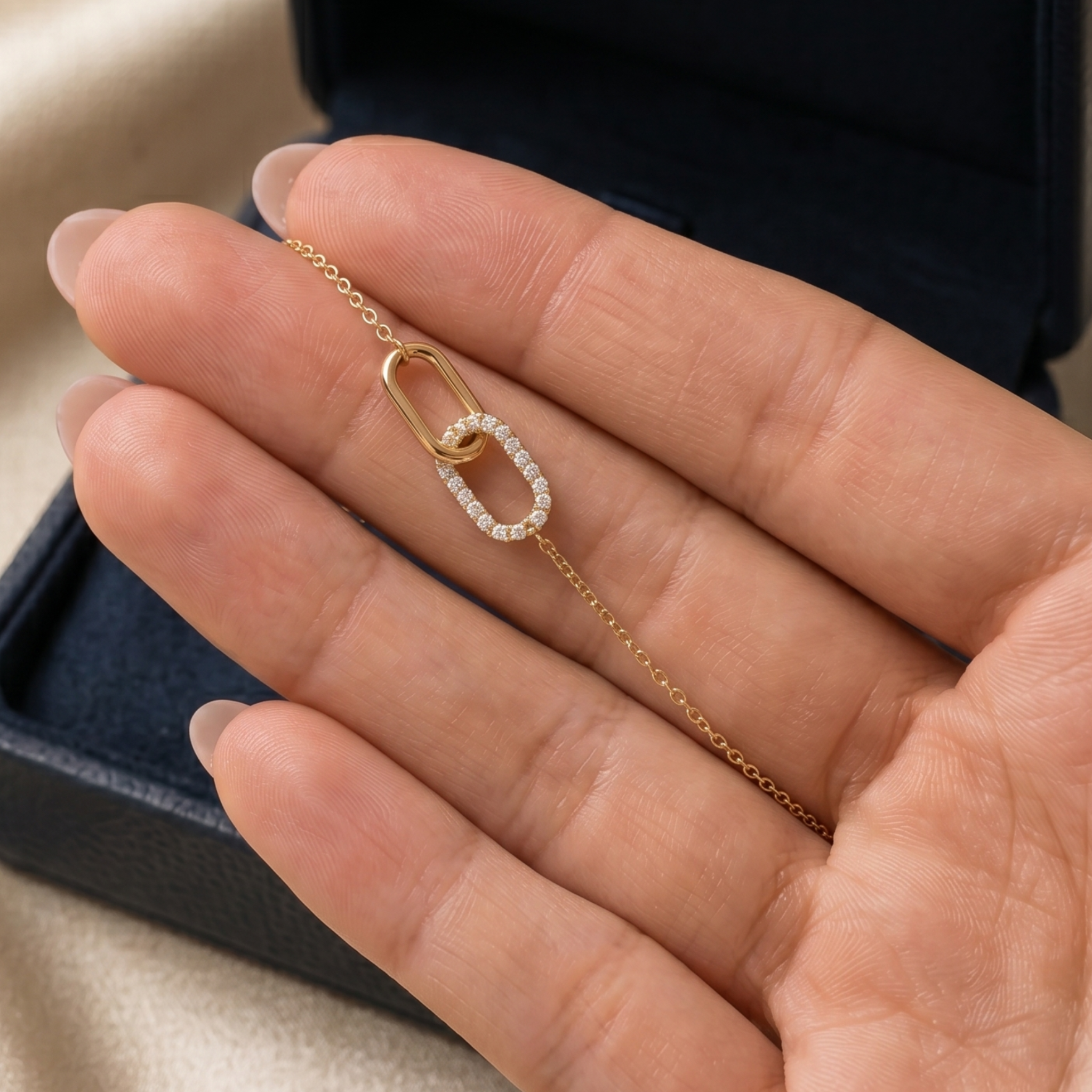 A delicate gold chain necklace with a double-link design, one side paved with small clear stones and the center linked with a gold ball, held on a person's hand. A dark jewelry box is visible in the background.