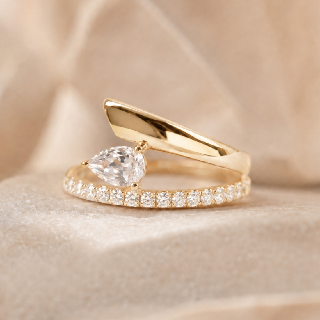 A gold ring featuring a pear-cut clear gemstone and a pavé band, displayed on a soft beige fabric background.