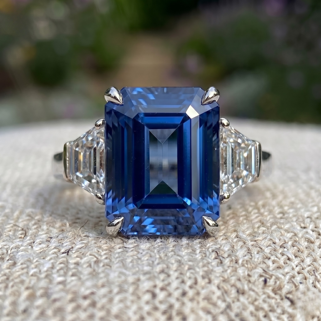 An emerald-cut blue sapphire ring with baguette side stones in a silver-toned setting, resting on a textured beige fabric with a blurred garden background.