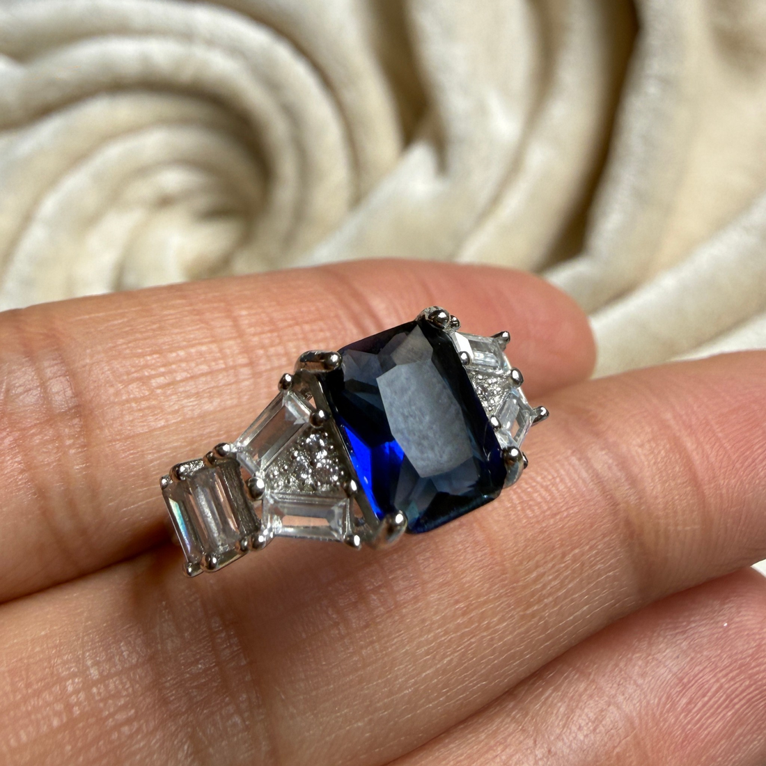 A silver-toned ring featuring a rectangular blue sapphire center stone and baguette-cut clear stones on either side, shown resting on a person's fingers against a soft cream fabric background.