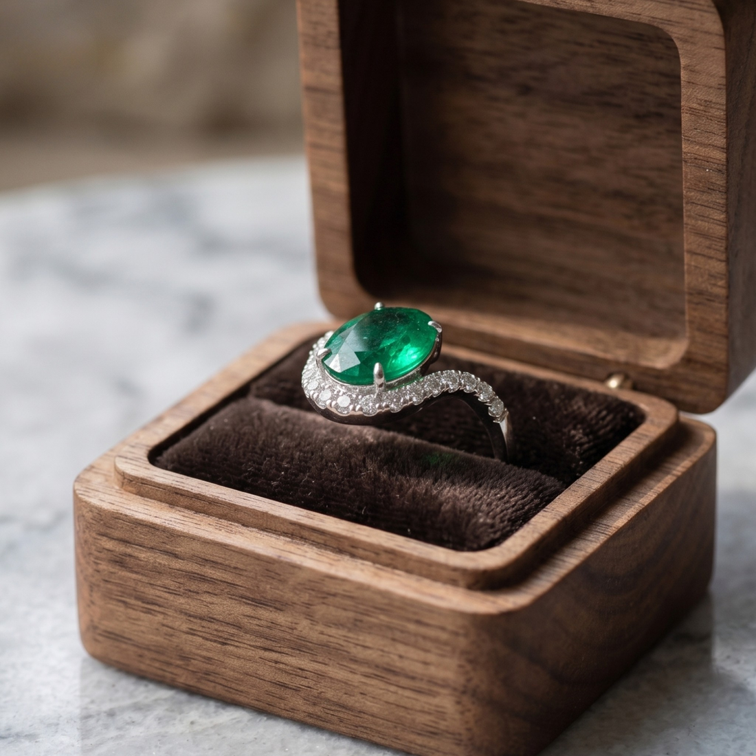 A silver ring with a large oval green emerald-cut center stone and small pavé-set stones, displayed in an open wooden jewelry box on a marble surface.