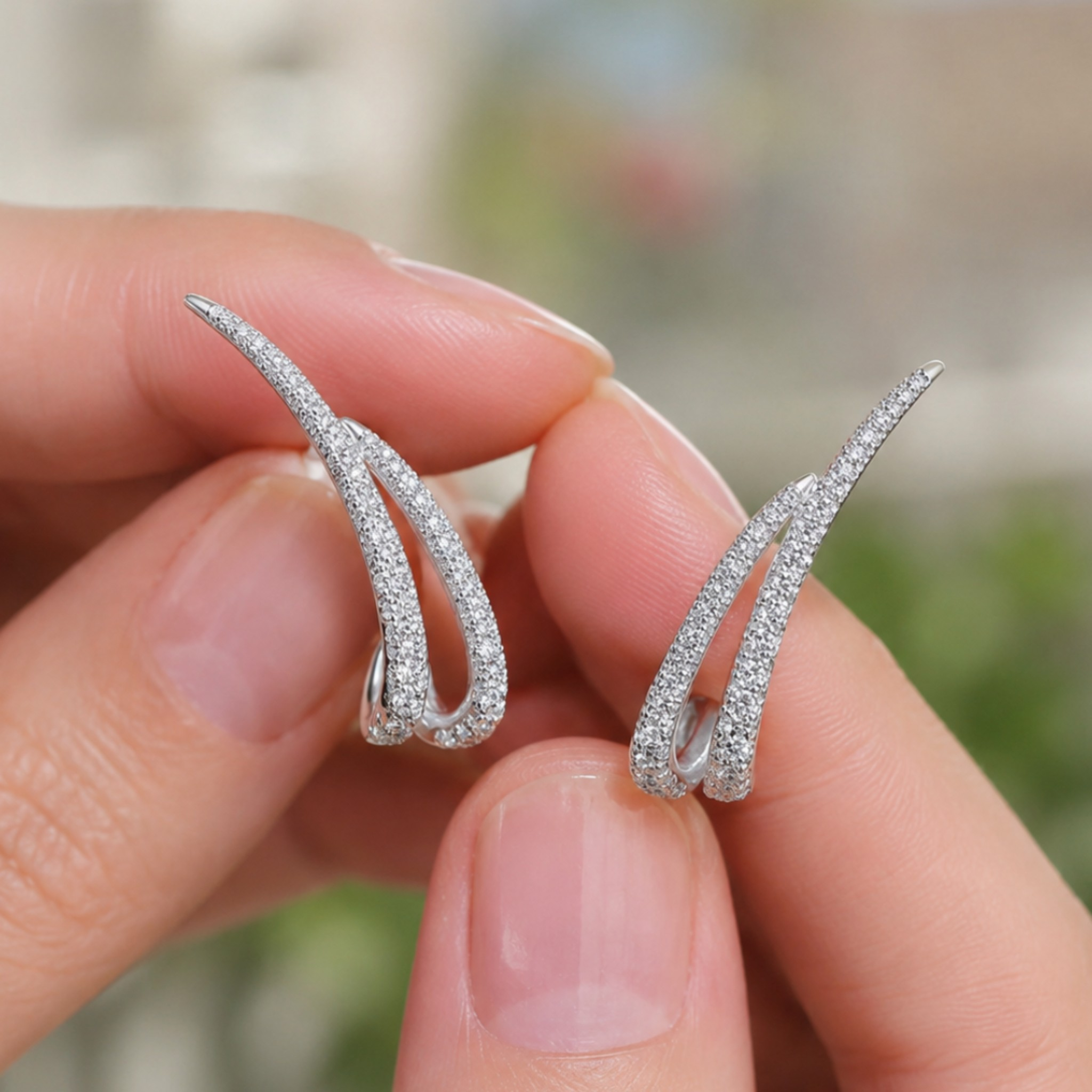 A pair of silver huggie hoop earrings paved with small clear stones, held between two fingers against a blurred outdoor background.