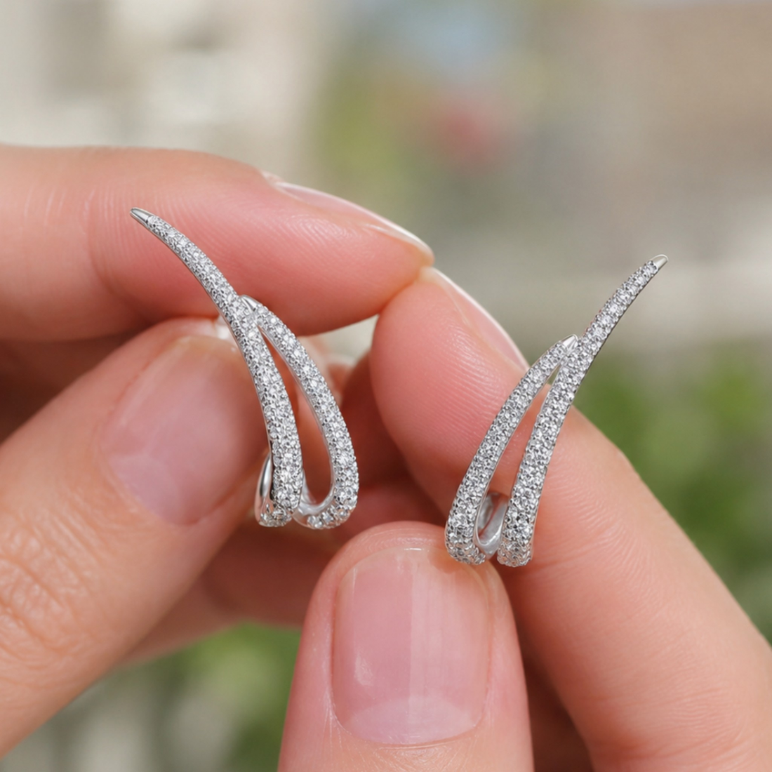 A pair of silver huggie hoop earrings paved with small clear stones, held between two fingers against a blurred outdoor background.