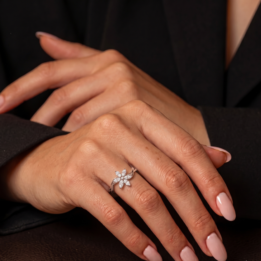 A silver-toned flower ring with small clear stones is worn on a woman's ring finger. The hands have pale pink manicured nails, and the person is wearing a black blazer.