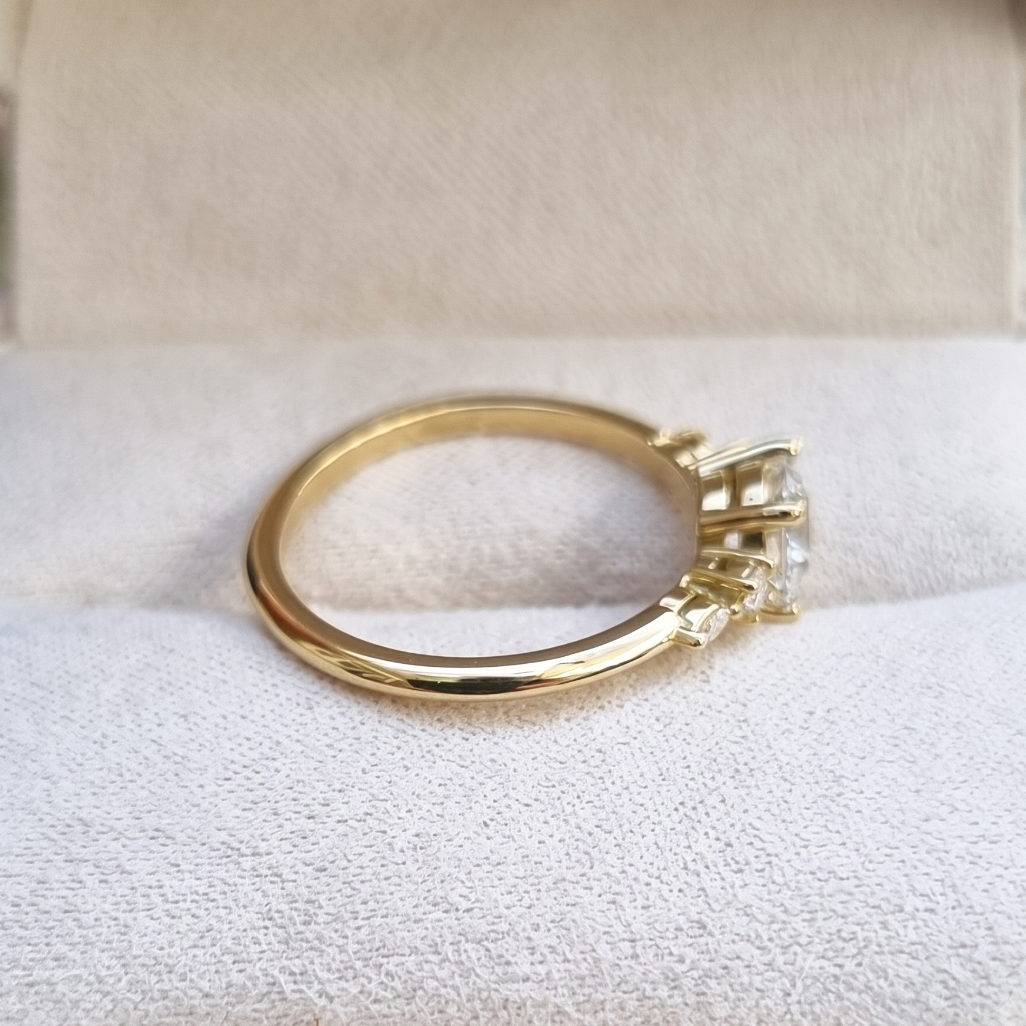 A yellow gold three-stone ring with clear gemstones, displayed on a textured white fabric surface inside a cream-colored jewelry box.