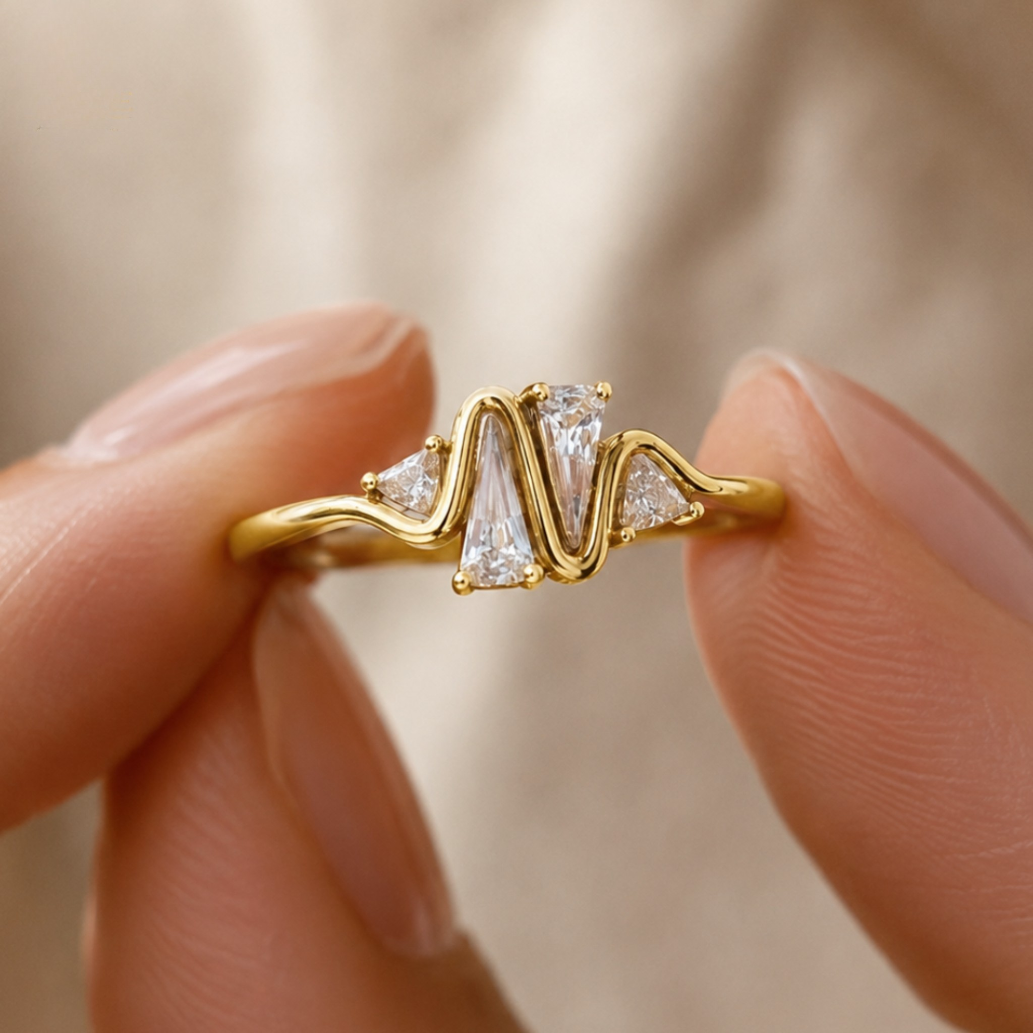A close-up of a gold ring with a wavy, sculptural band and clear baguette and round-cut stones, held between two fingers against a soft beige fabric background.