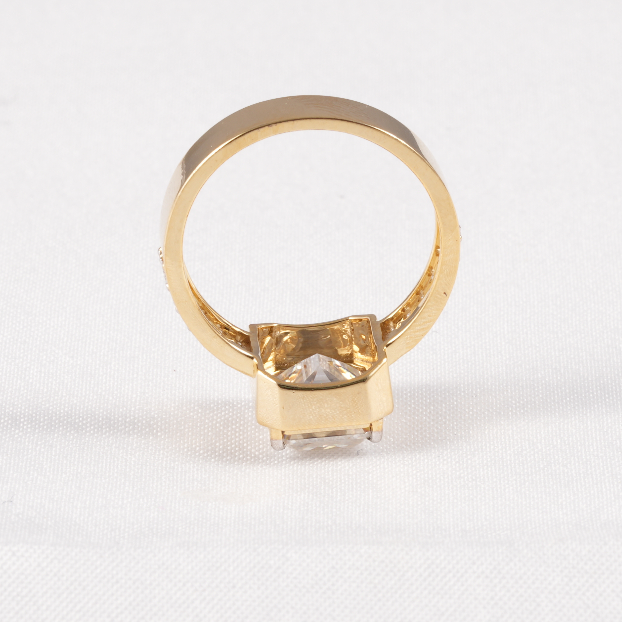 A gold ring with a square bezel setting holding a clear, faceted gemstone, shown from the back on a textured white fabric background.