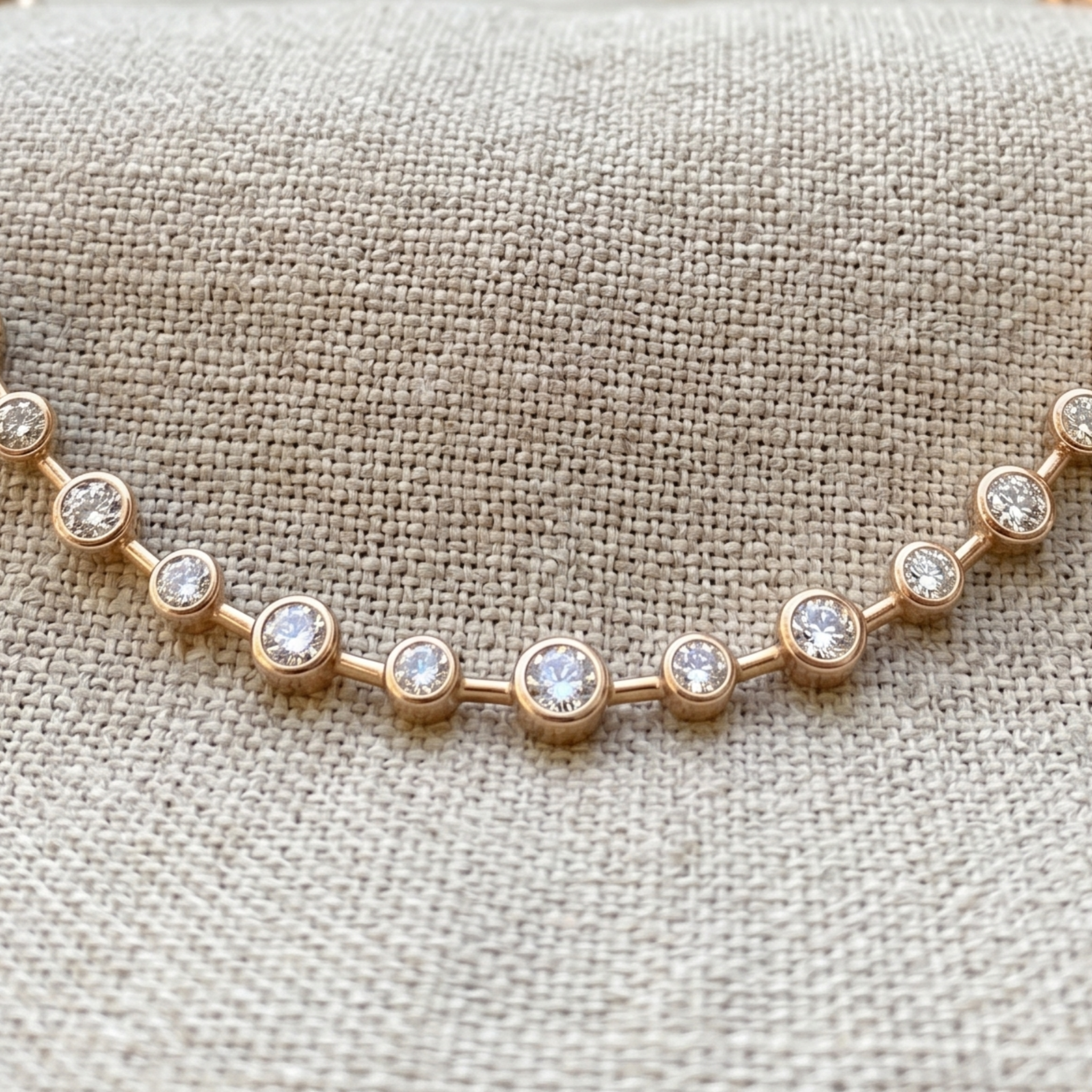 A close-up of a delicate rose gold bracelet with round bezel-set clear stones, displayed on a textured beige linen fabric background.