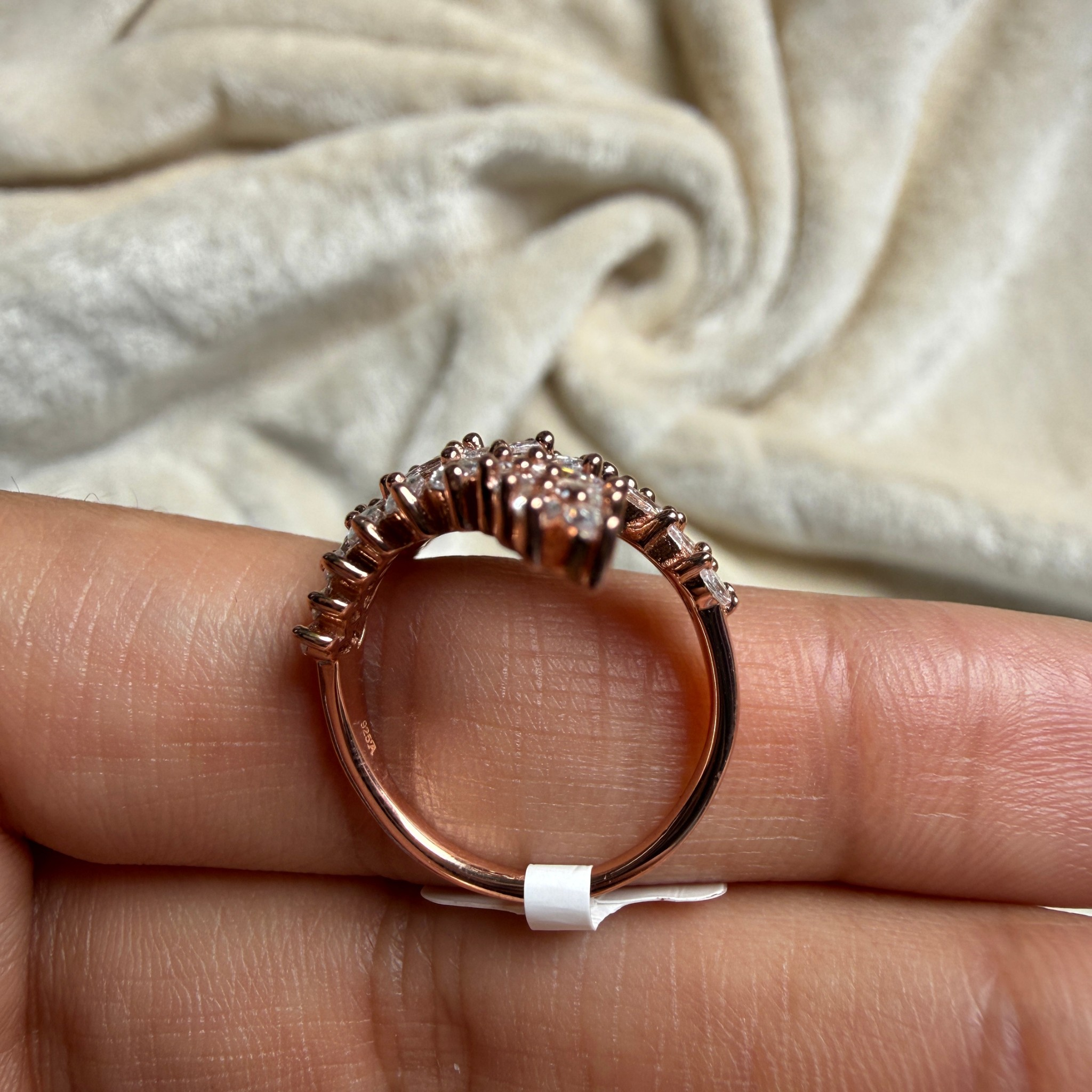 A rose gold ring with a textured, scalloped edge and small clear stones, held on a person's fingers against a soft beige fabric background.