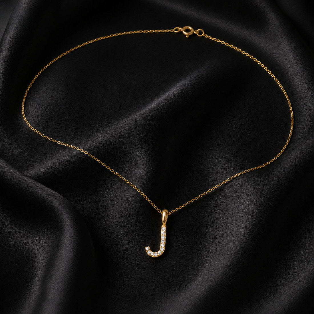 A delicate gold chain necklace with a small letter 'J' pendant adorned with clear pavé stones, displayed on a draped black satin fabric background.