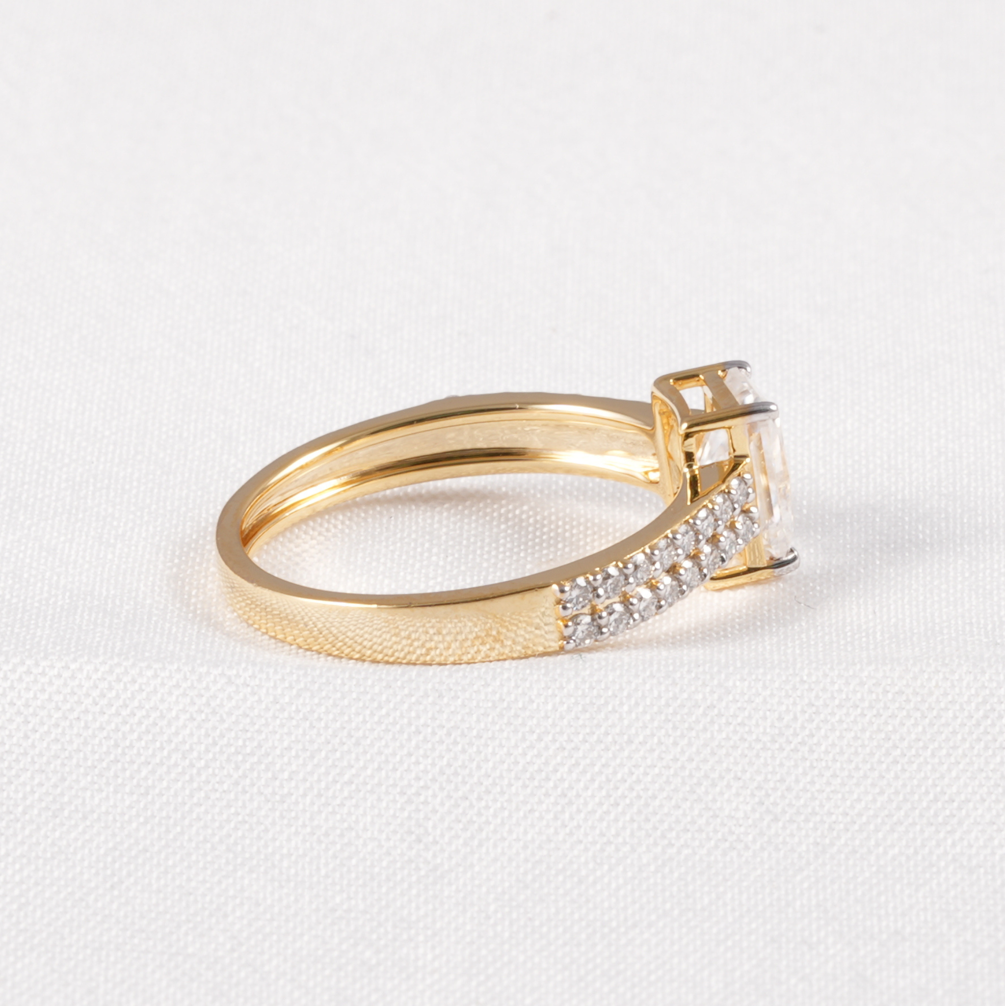 A yellow gold ring with a textured band, a pavé-set row of clear stones, and a larger central clear gemstone, photographed on a white textured fabric background.