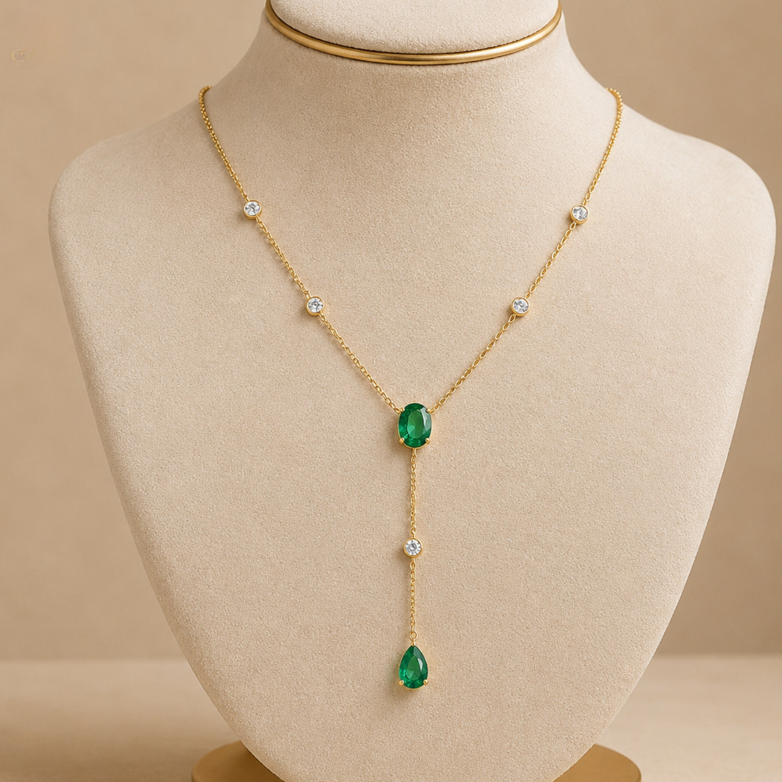 A gold Y-necklace with a large oval green emerald at the center and a smaller teardrop green emerald at the bottom, accented with small clear round stones, displayed on a beige jewelry bust.