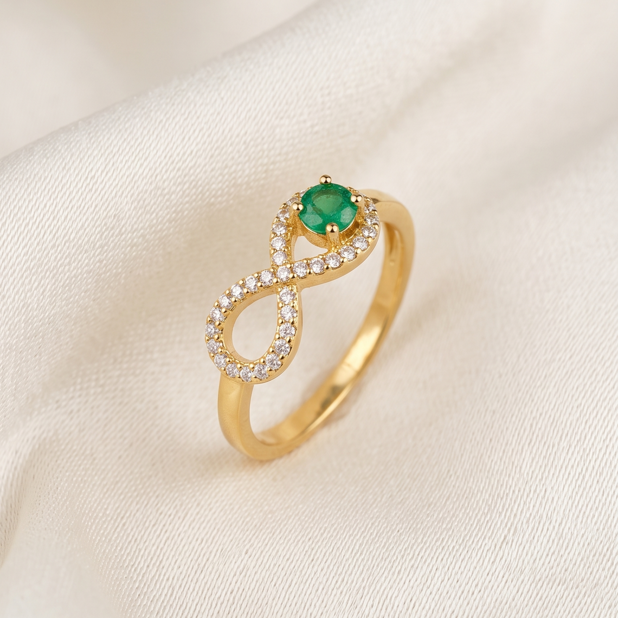 A gold infinity-style ring featuring a round green gemstone at the center and a halo of small clear stones along the upper loop, displayed on a soft cream fabric background.