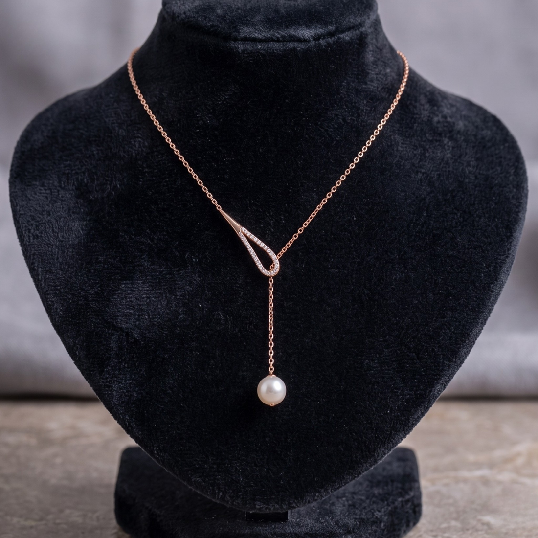 A delicate rose gold necklace with a Y-shaped design, featuring a small diamond-like accent at the top and a single dangling pearl at the bottom, displayed on a black velvet jewelry bust.