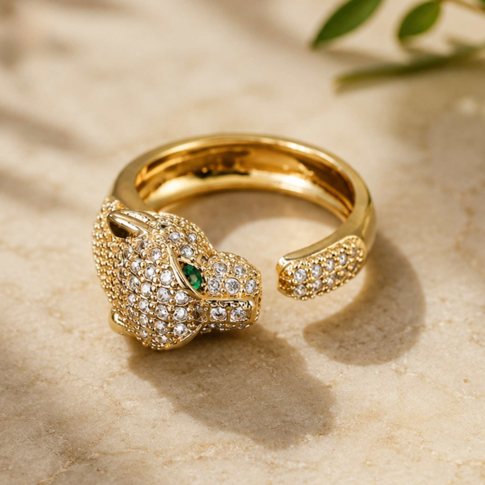 A gold-toned open band ring featuring a stylized animal head pavé-set with clear stones and a small green stone accent, photographed on a light beige textured surface with soft shadows and blurred green leaves in the background.