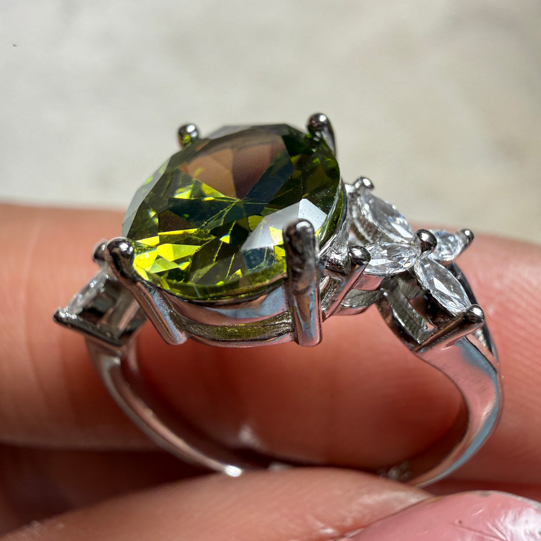 A silver ring featuring a large oval green gemstone with marquise-cut side stones, shown being held between fingers.